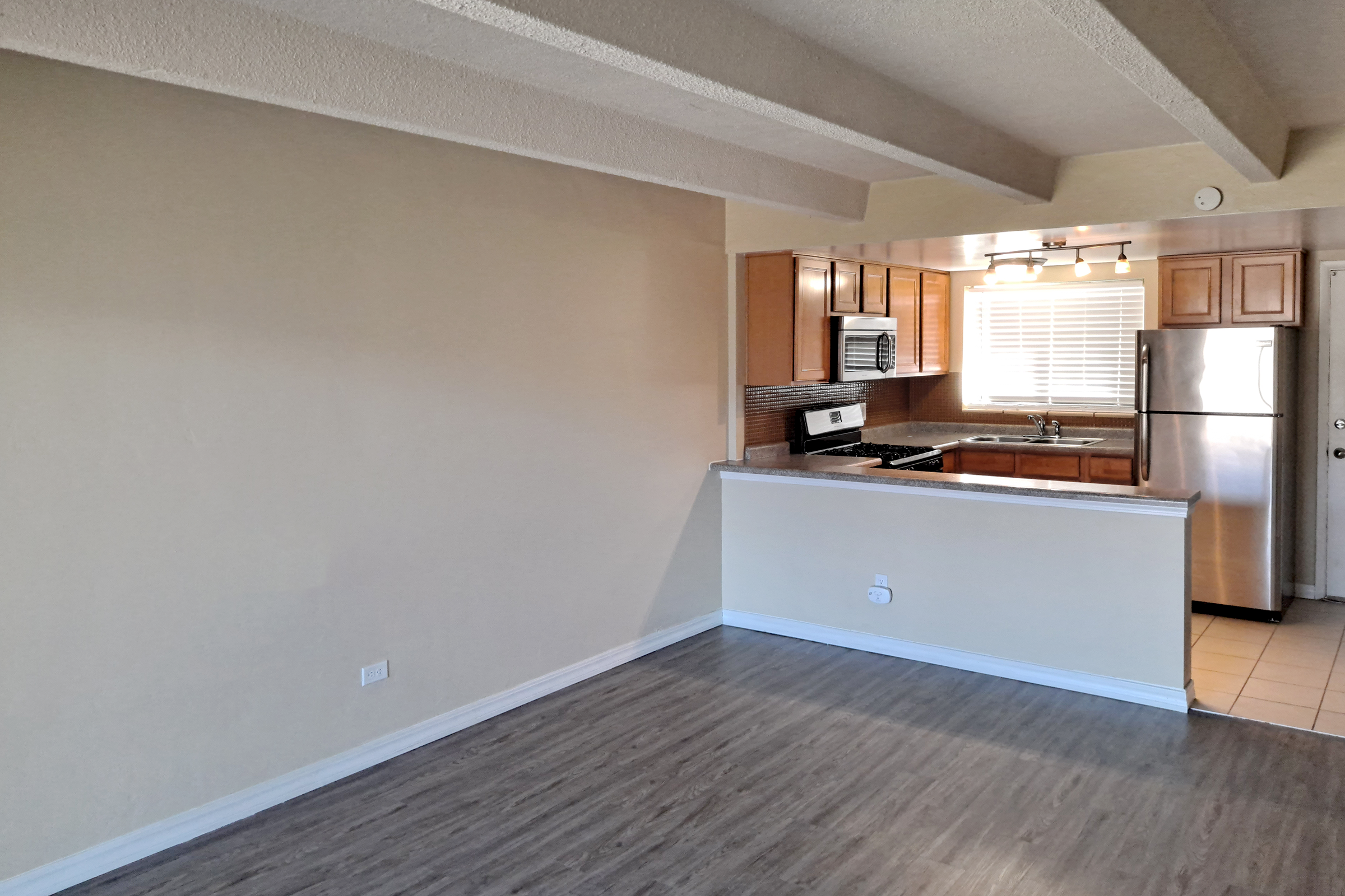 An open-concept living space featuring a beige wall and wood-like flooring. The kitchen is visible with wooden cabinets, stainless steel appliances, and a window providing natural light. A small bar area separates the kitchen from the living room, creating an inviting atmosphere.