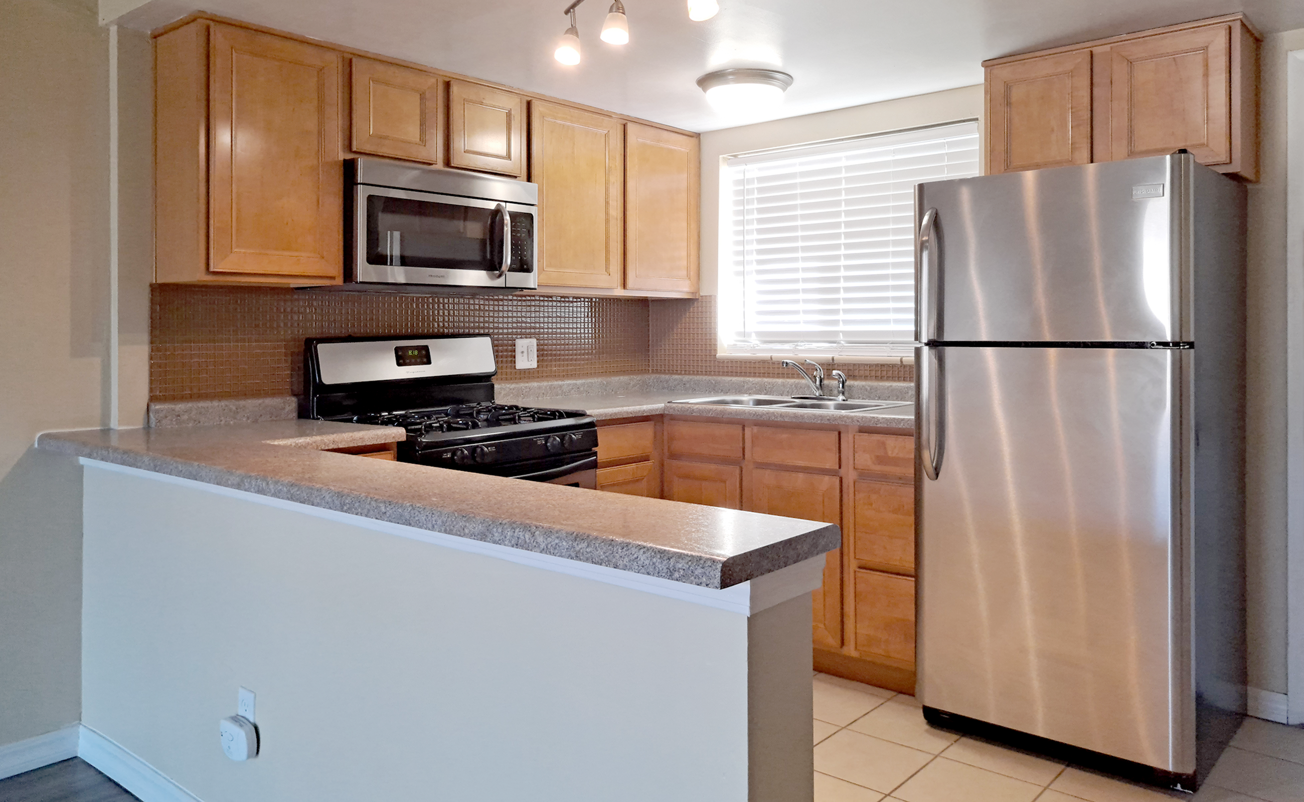 A modern kitchen featuring wooden cabinetry, stainless steel appliances including a microwave, gas stove, and refrigerator. The room has a light-colored countertop, tiled flooring, and a window allowing natural light. Bright overhead lights enhance the space's functionality and appeal.