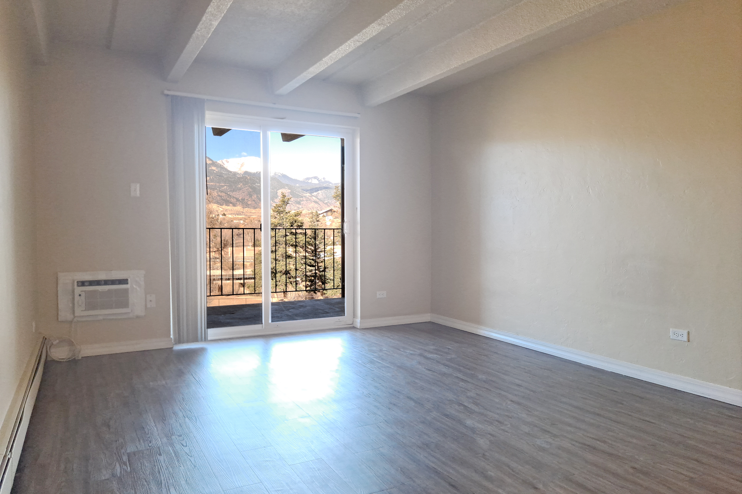 A spacious, empty room with light-colored walls and wooden floors. A sliding glass door leads to a balcony with a view of distant mountains and trees. The room is well-lit with natural light, and there's an air conditioning unit on one wall. The ceiling features exposed beams for added character.