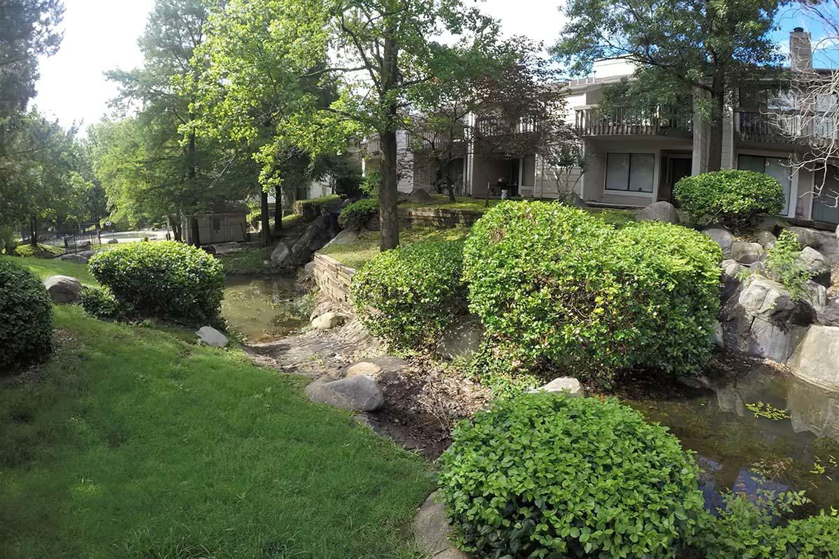 A peaceful outdoor scene featuring a serene pond surrounded by lush greenery and neatly trimmed bushes. In the background, there are apartment buildings partially obscured by trees, creating a tranquil and inviting atmosphere. Sunlight filters through the leaves, enhancing the natural beauty of the setting.
