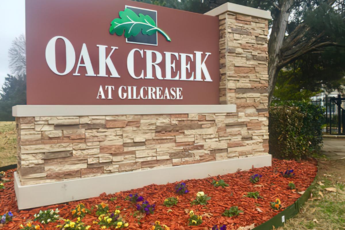 Sign for Oak Creek at Gilcrease, featuring the name in large, white letters against a brown background, with a green leaf graphic above it. Surrounding the sign are flower beds with colorful blooms and decorative mulch, set in a landscaped area with trees and shrubbery in the background.