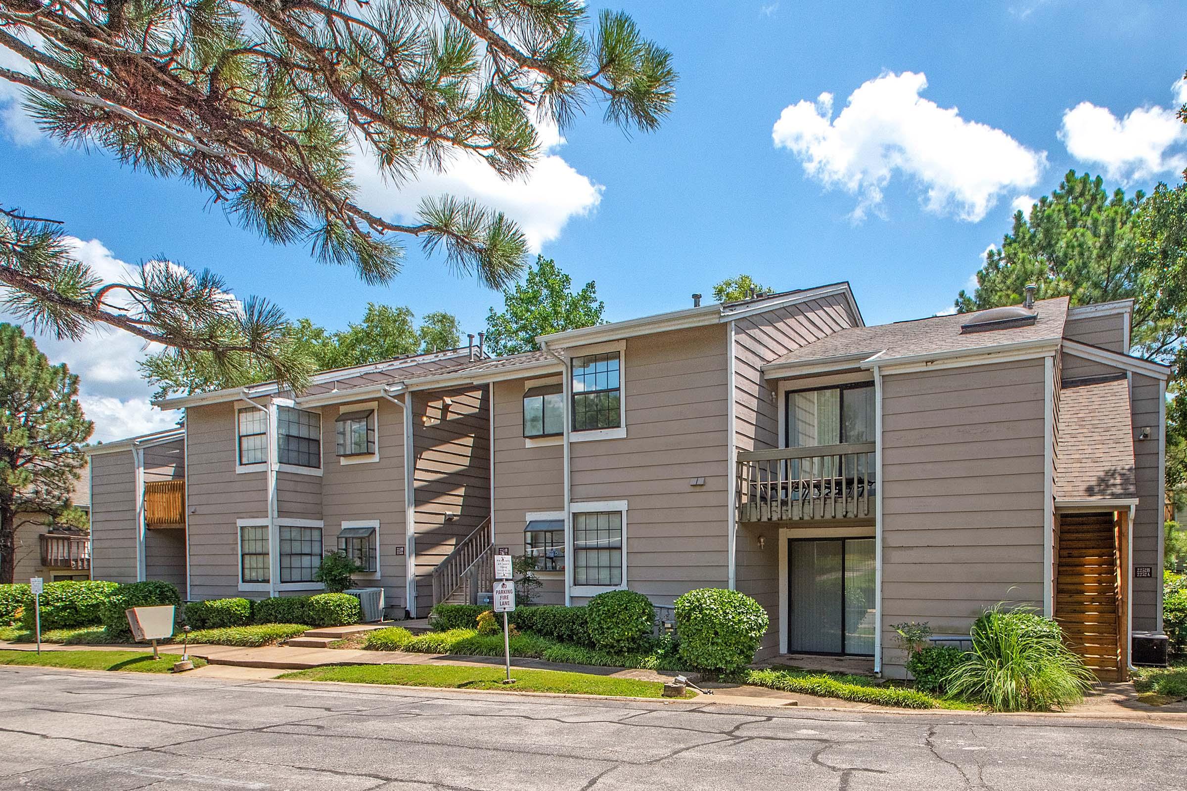 Two-story apartment building with a light brown exterior, surrounded by lush greenery and trees. The facade features balconies and staircases, with a clear blue sky and some fluffy clouds in the background. A paved road runs in front of the building, enhancing the residential neighborhood feel.