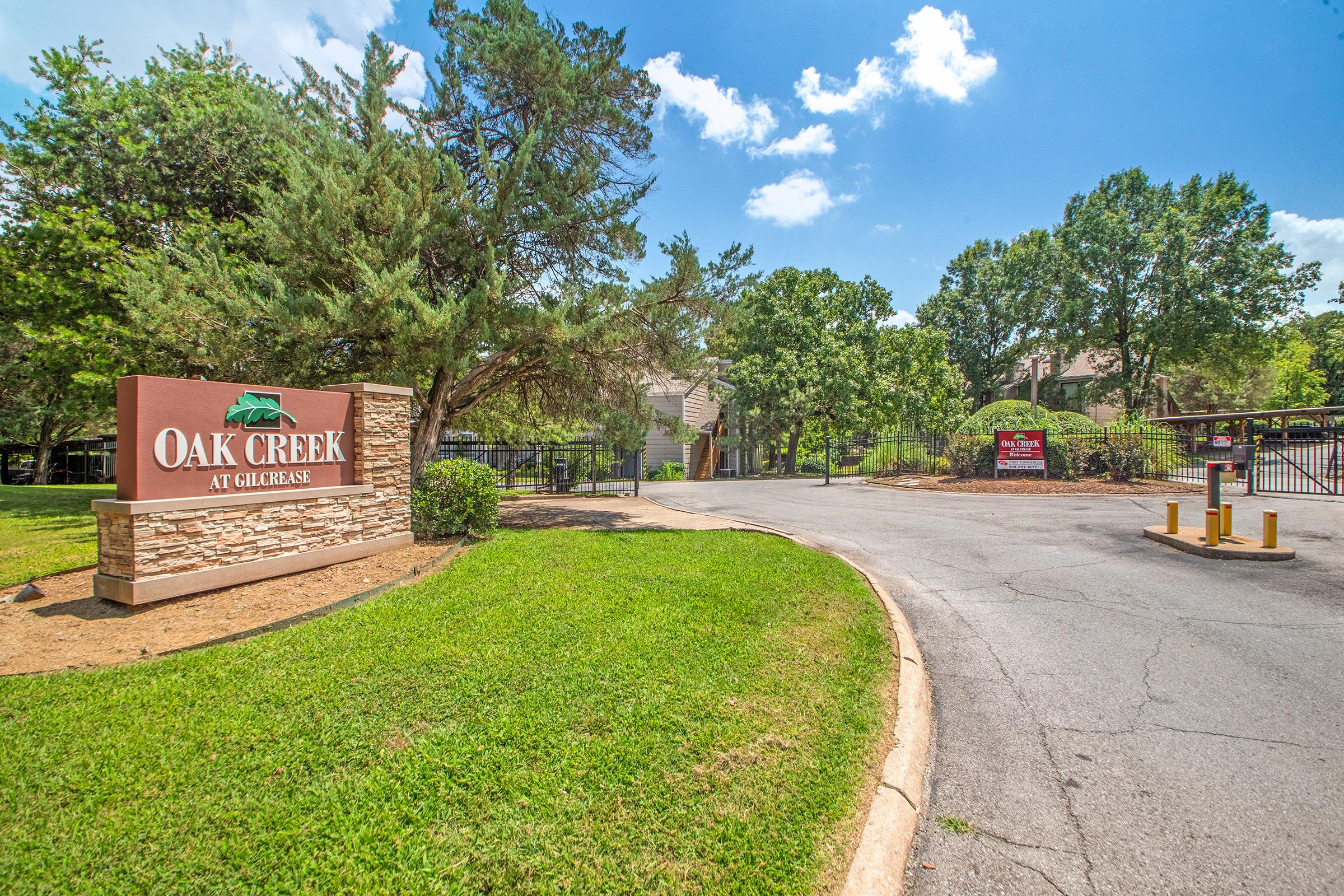 A view of the entrance to Oak Creek at Galgari, featuring a welcoming sign among lush greenery. The scene includes trees, manicured grass, and a gated entryway, with blue skies above.
