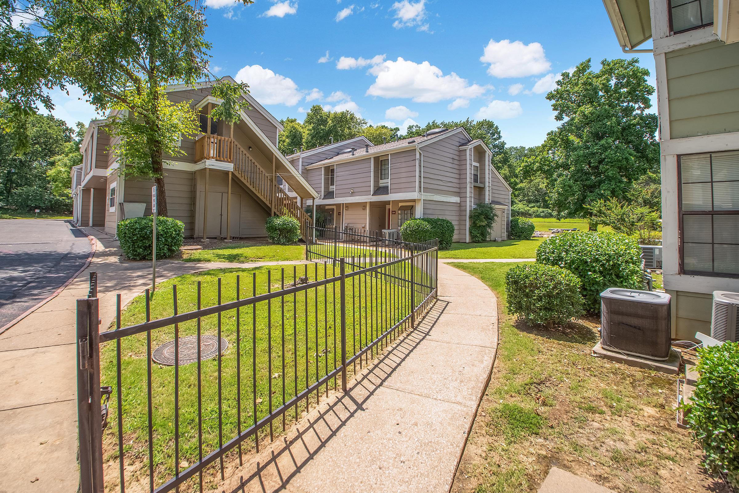 A pathway lined with green bushes leads to two multi-story apartment buildings situated in a sunny landscape. A wrought-iron fence encloses the pathway, and there are trees in the background under a bright blue sky dotted with fluffy white clouds.