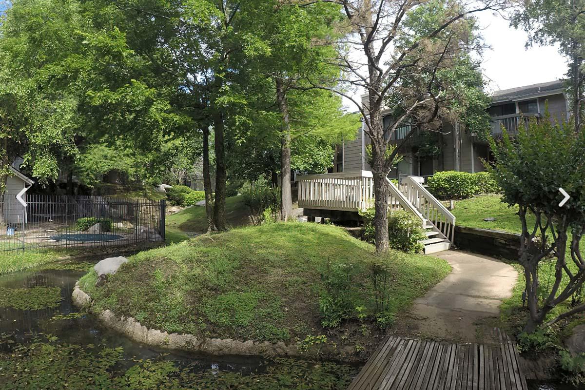 A peaceful outdoor scene featuring lush greenery, a small hill, and a wooden walkway leading to a bridge. In the background, a building is partially visible, and there is a fenced area, likely a small animal enclosure, next to a calm water feature with some vegetation.