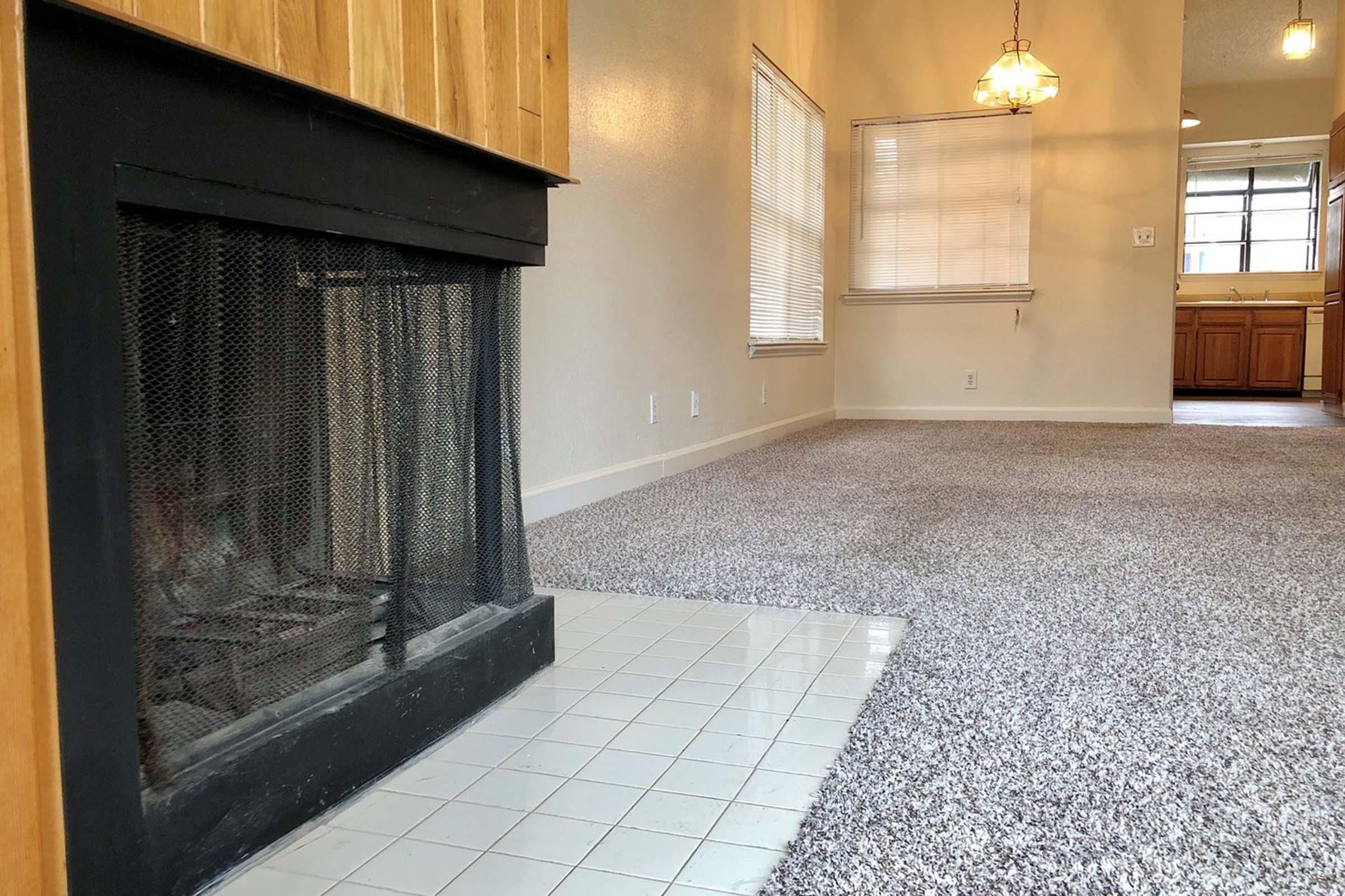 A cozy living room featuring a tile fireplace with a metal screen, plush gray carpet, and warm wooden accents. Large windows allow natural light, and an open layout leads to a kitchen with wooden cabinetry visible in the background. Two pendant lights hang from the ceiling, adding a welcoming touch.
