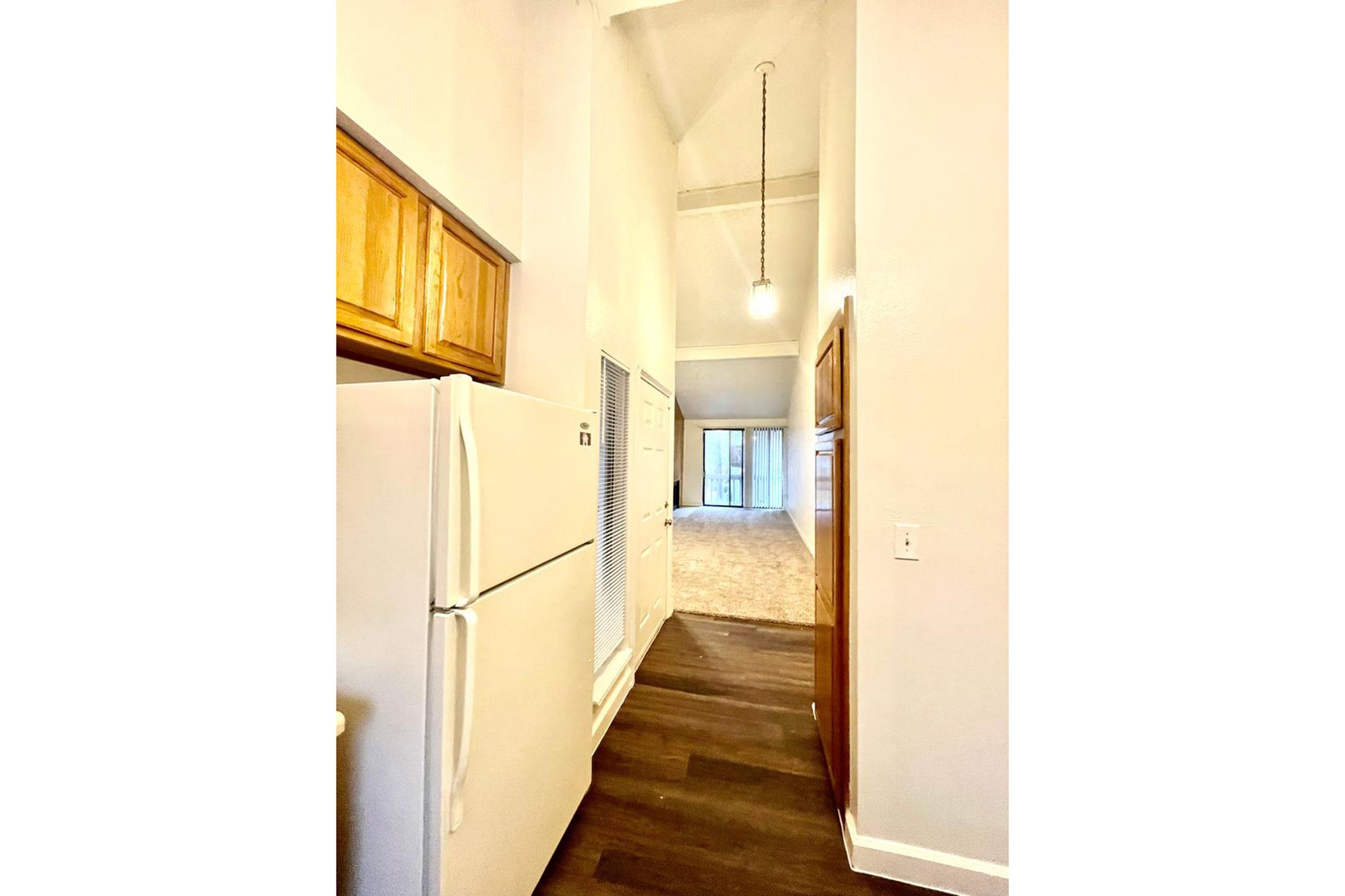Narrow kitchen hallway featuring a white refrigerator on the left, light wood cabinetry above, and a light fixture hanging from a high ceiling. The hallway leads to a spacious living area with large windows visible in the background, allowing natural light to illuminate the space.