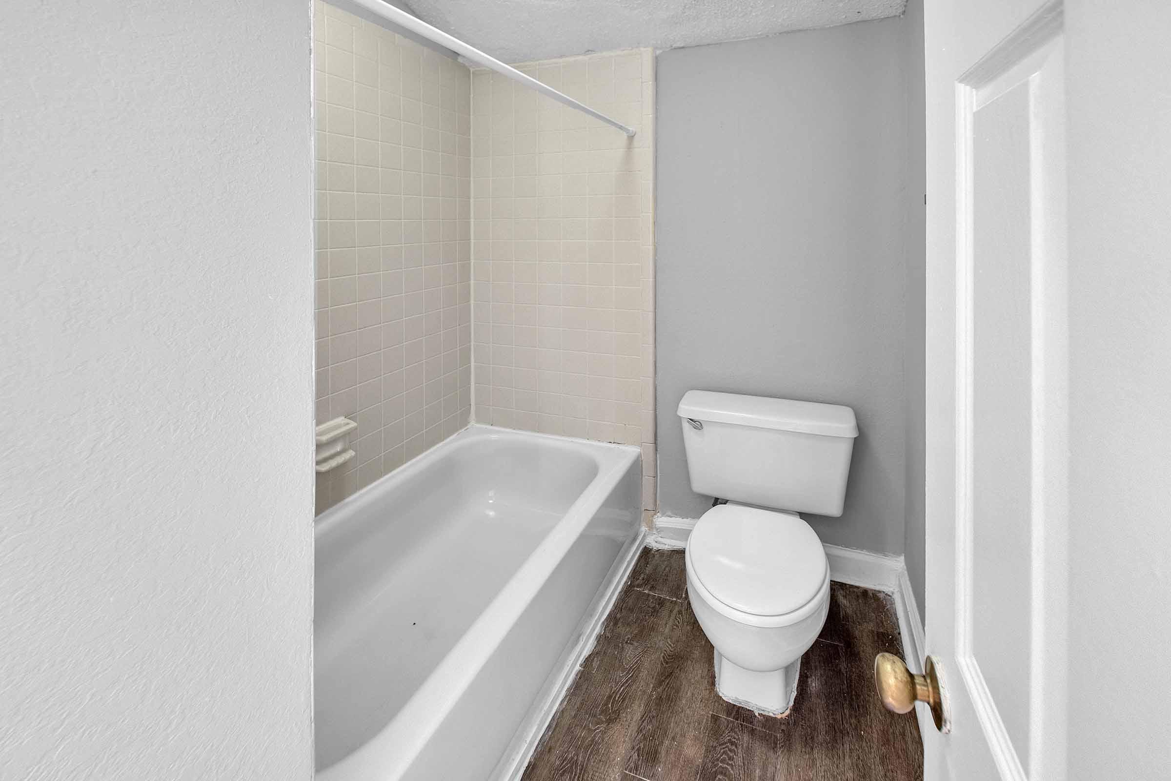A clean, simple bathroom featuring a bathtub with a shower rod, a toilet, and a painted gray wall. The floor is covered with brown laminate, and the bathroom has neutral-colored tiles along the bathtub area.