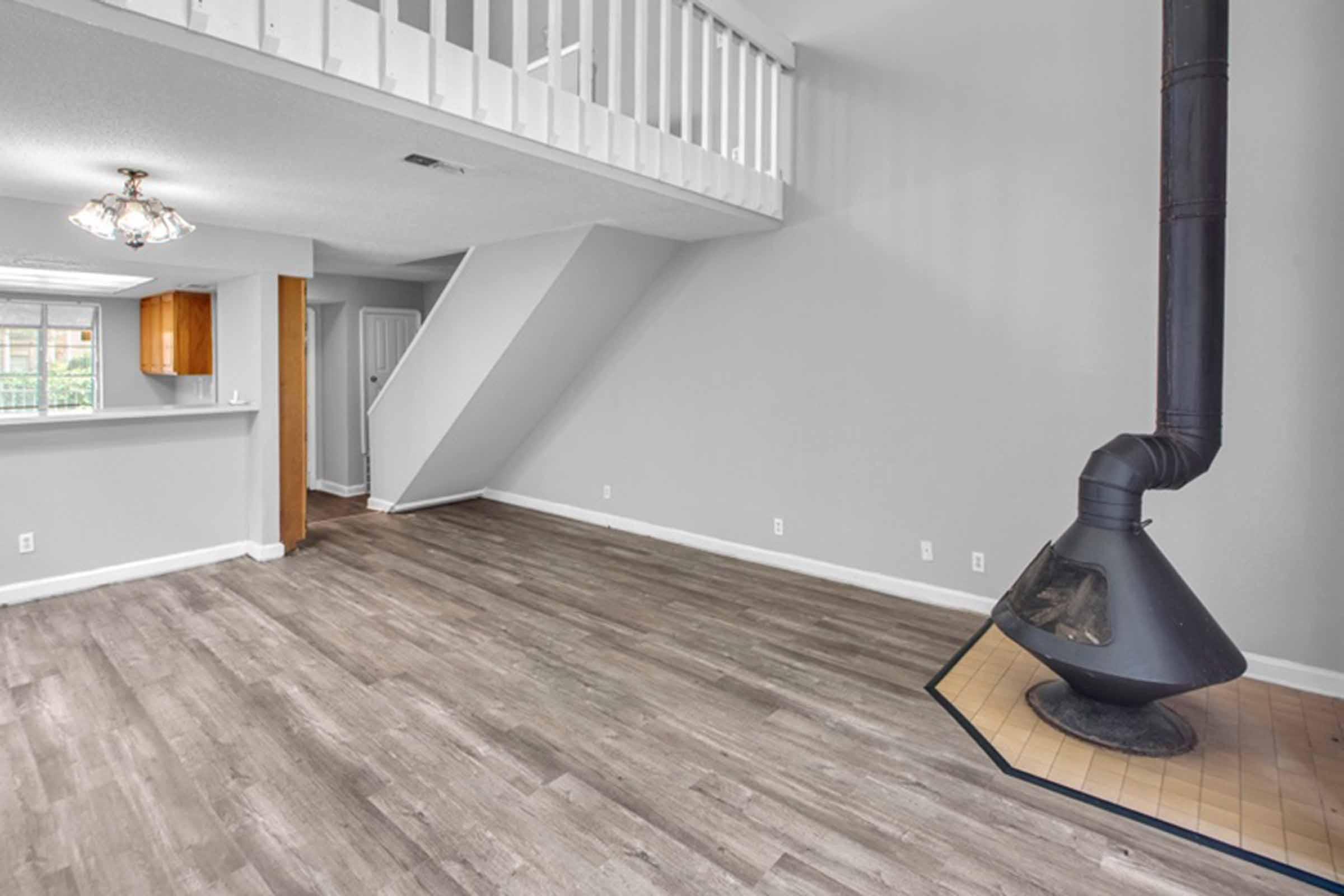 Interior view of a spacious living room featuring light gray walls and laminate flooring. There is a modern, black wood stove in the corner and a staircase leading to an upper level. A large window provides natural light, and there's an open layout connecting to the kitchen and dining area.