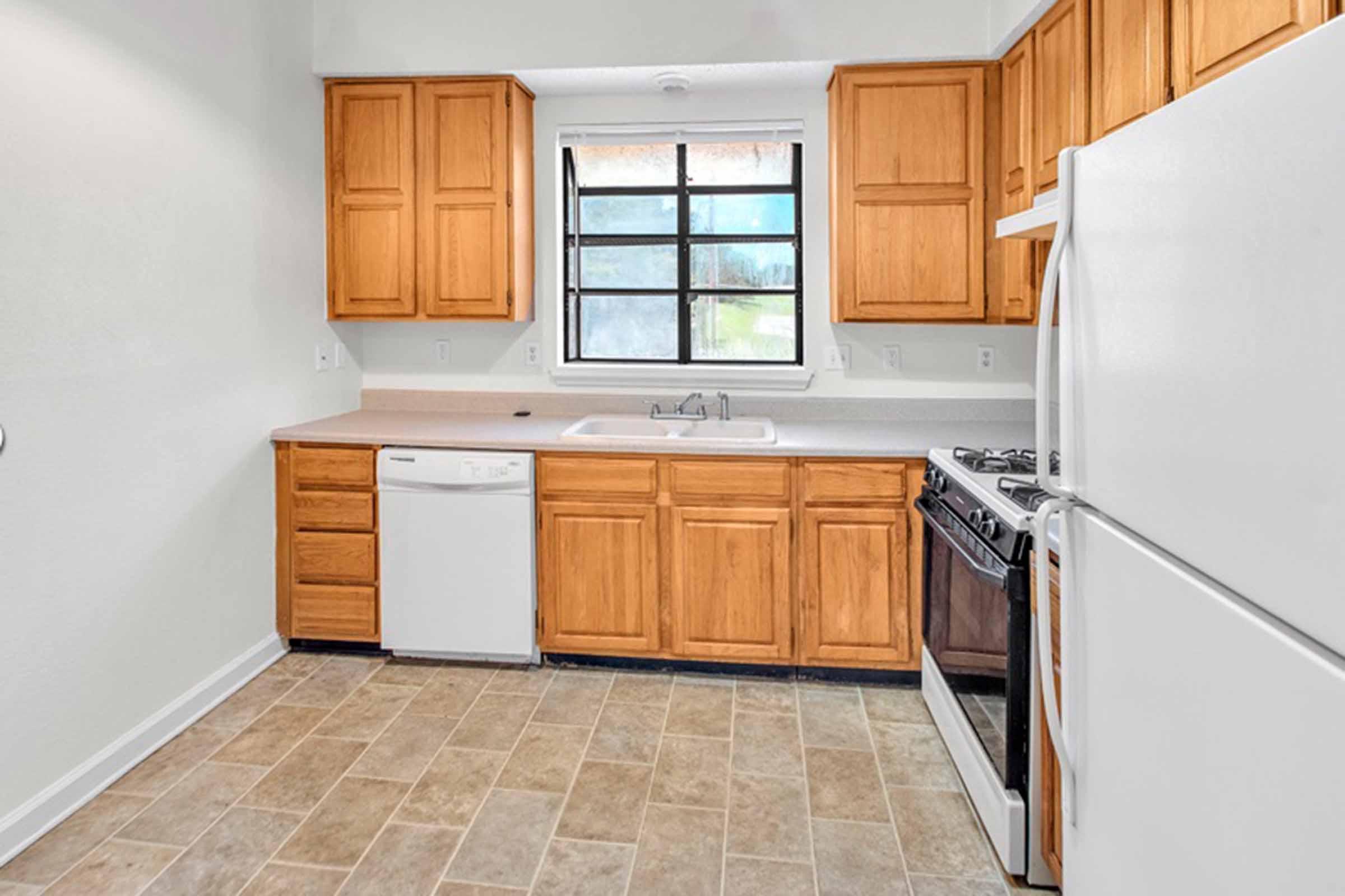 A modern kitchen featuring wooden cabinets, a white dishwasher, a stainless steel gas stove, and a sink under a window. The floor is tiled with light-colored squares, creating a spacious and clean appearance.