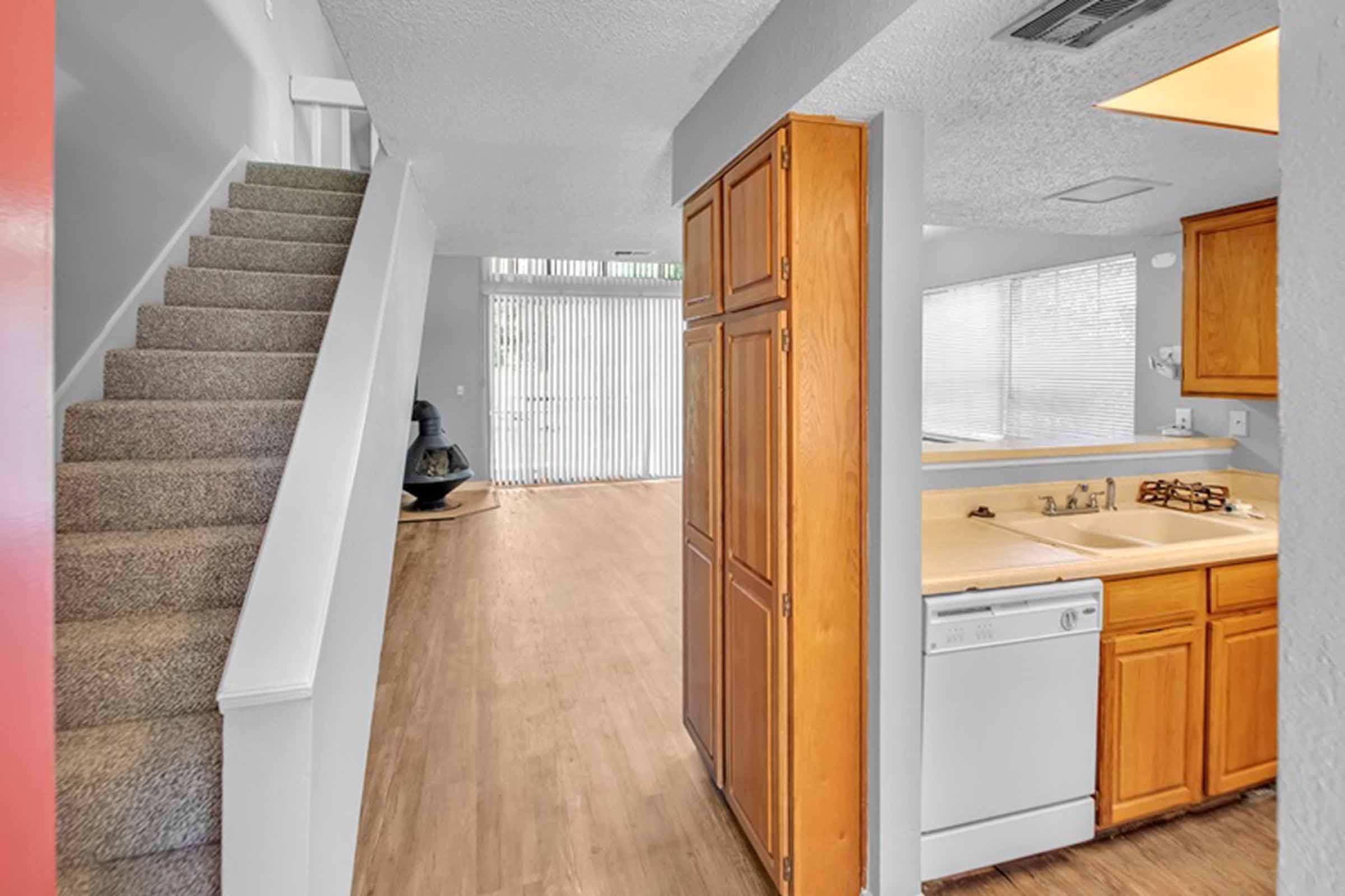 Interior view of a home featuring a staircase on the left side, an open kitchen area with wooden cabinets, and a dishwasher. A spacious living area is visible at the back with sliding glass doors leading outside, allowing natural light to enter. The flooring is a light wood finish, creating a warm ambiance.