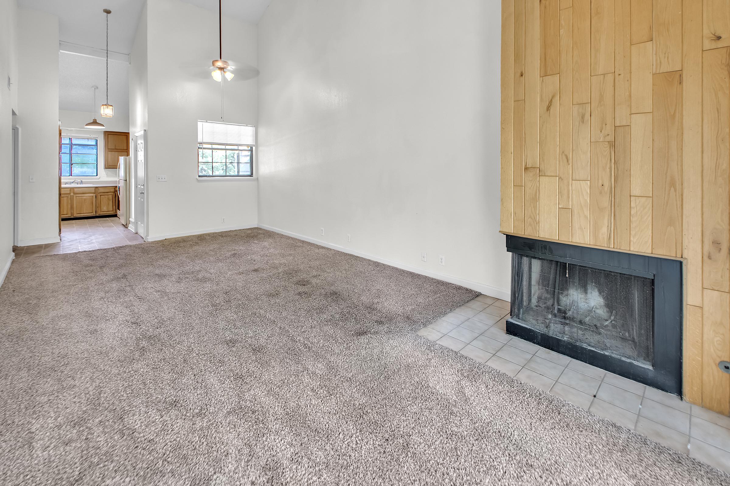 Spacious living area with high ceilings, beige carpet, and a wooden accent wall featuring a fireplace. Natural light streams in through a window, with a view of a kitchen area in the background. The room has a clean, open feel, ideal for various interior design options.