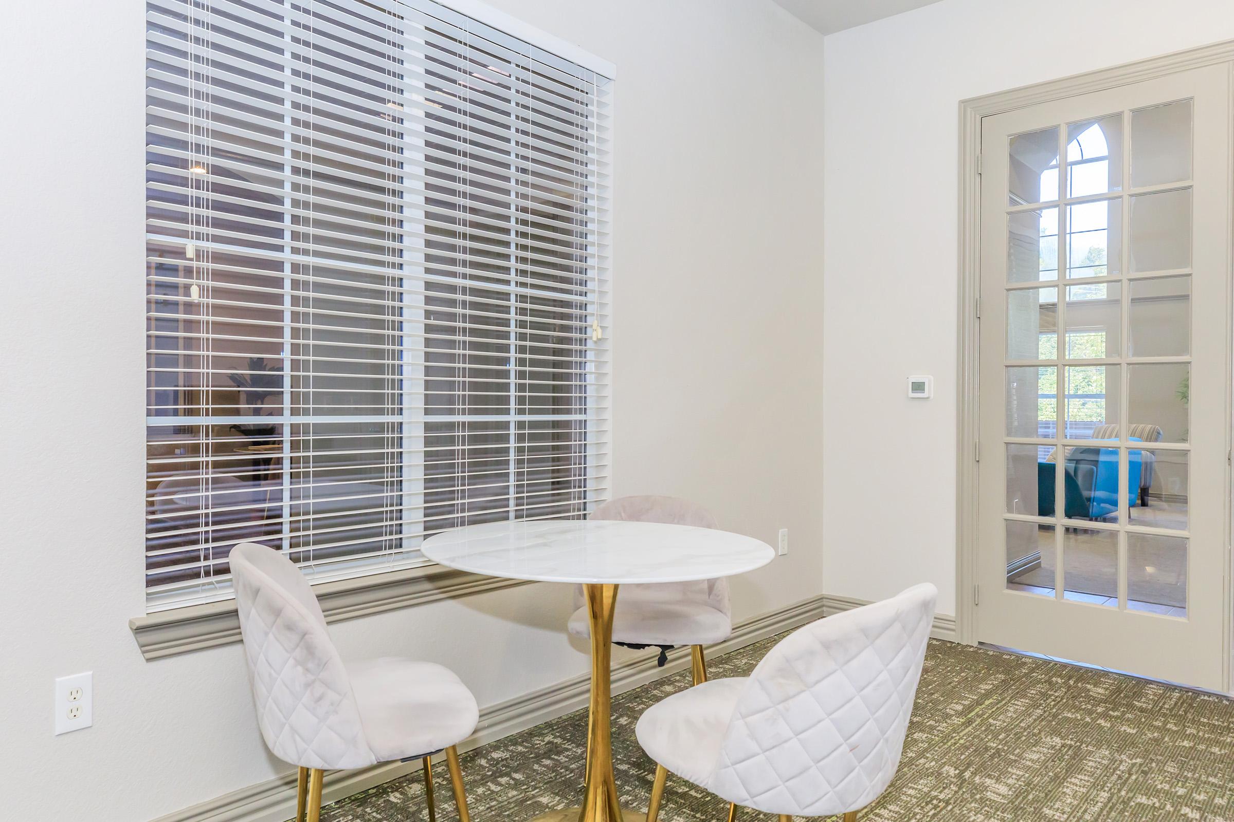 A small dining area featuring a round white table with a shiny gold base, accompanied by two plush, light-colored chairs. The background includes a window with white blinds and a door with glass panels leading to another room. The flooring is covered with a textured carpet.