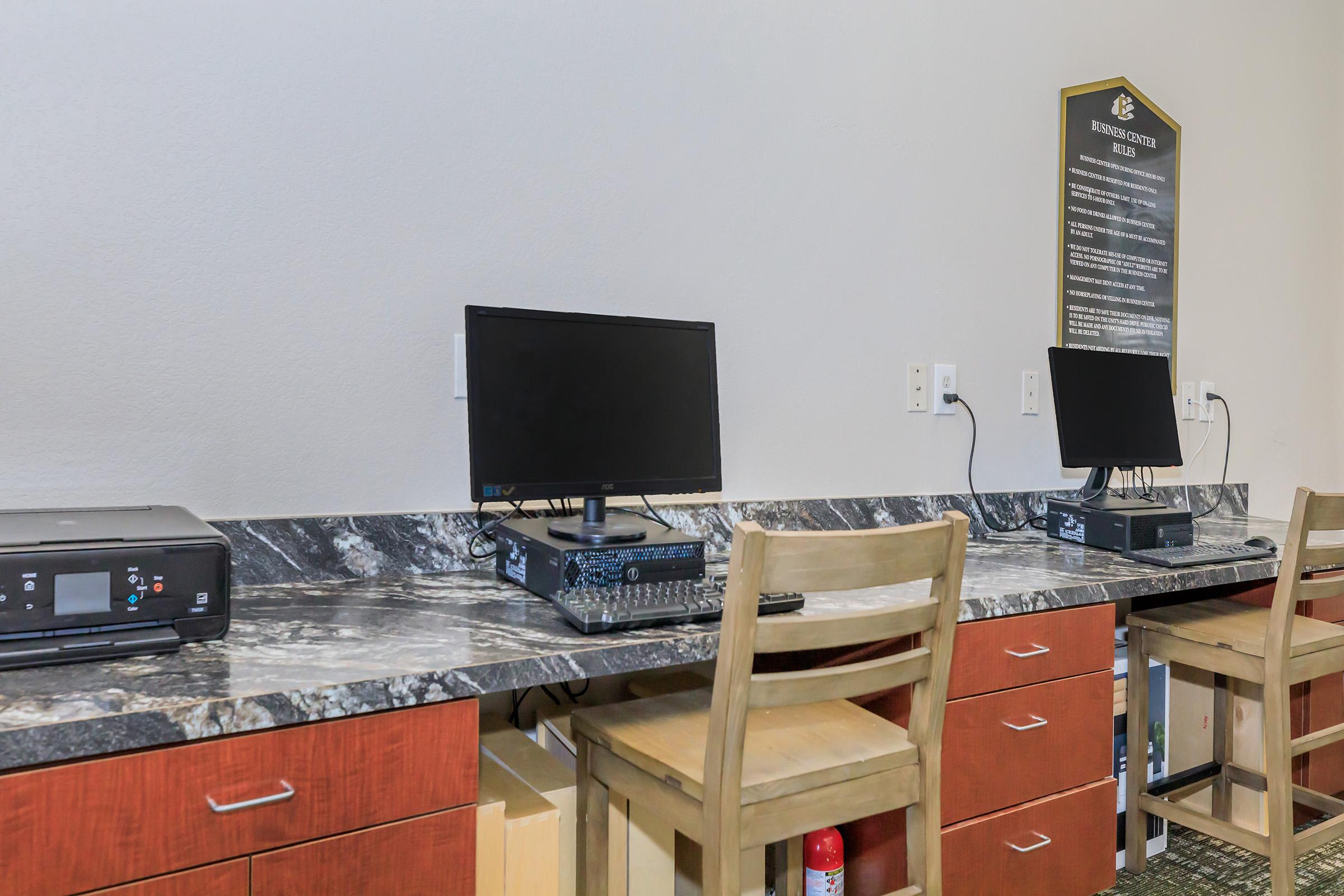 A small business center featuring a dark countertop with a desktop computer and printer. Two wooden chairs are positioned at the counter, with a wall-mounted information board in the background. The environment is well-lit and minimalistic, suitable for work or printing tasks.