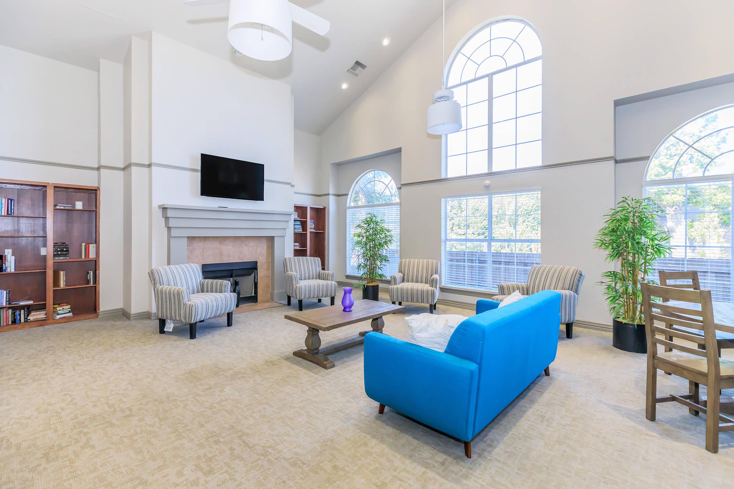 A modern, well-lit living room featuring a blue couch, two striped armchairs, and a wooden coffee table. There is a fireplace with a TV mounted above it and large windows that allow natural light to flood the space. Bookshelves with various books are visible on one side, as well as decorative plants adding a touch of greenery.