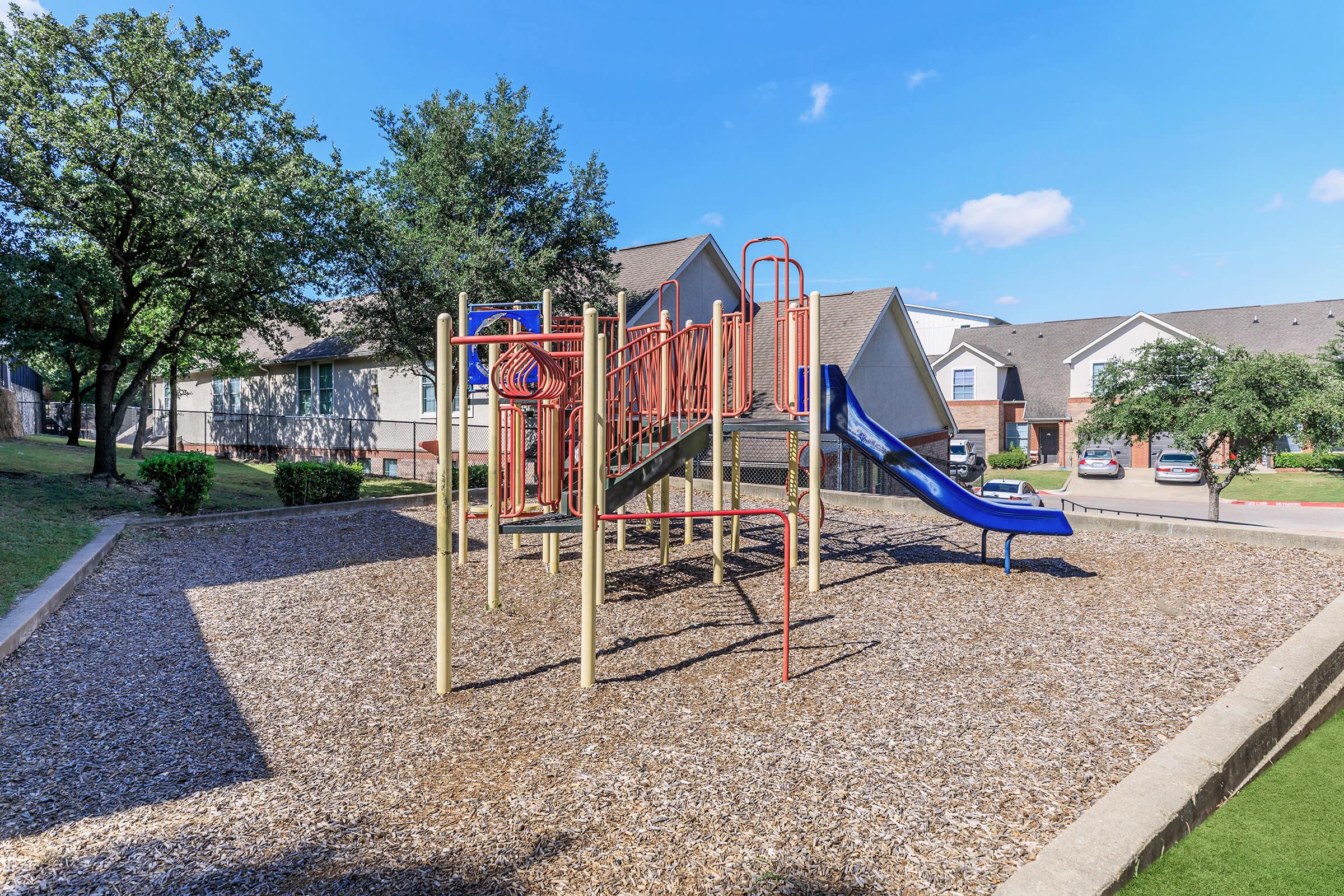 A playground featuring a climbing structure with a slide, surrounded by tan playground mulch and grassy areas. There are trees in the background, and several residential buildings are visible nearby under a clear blue sky.