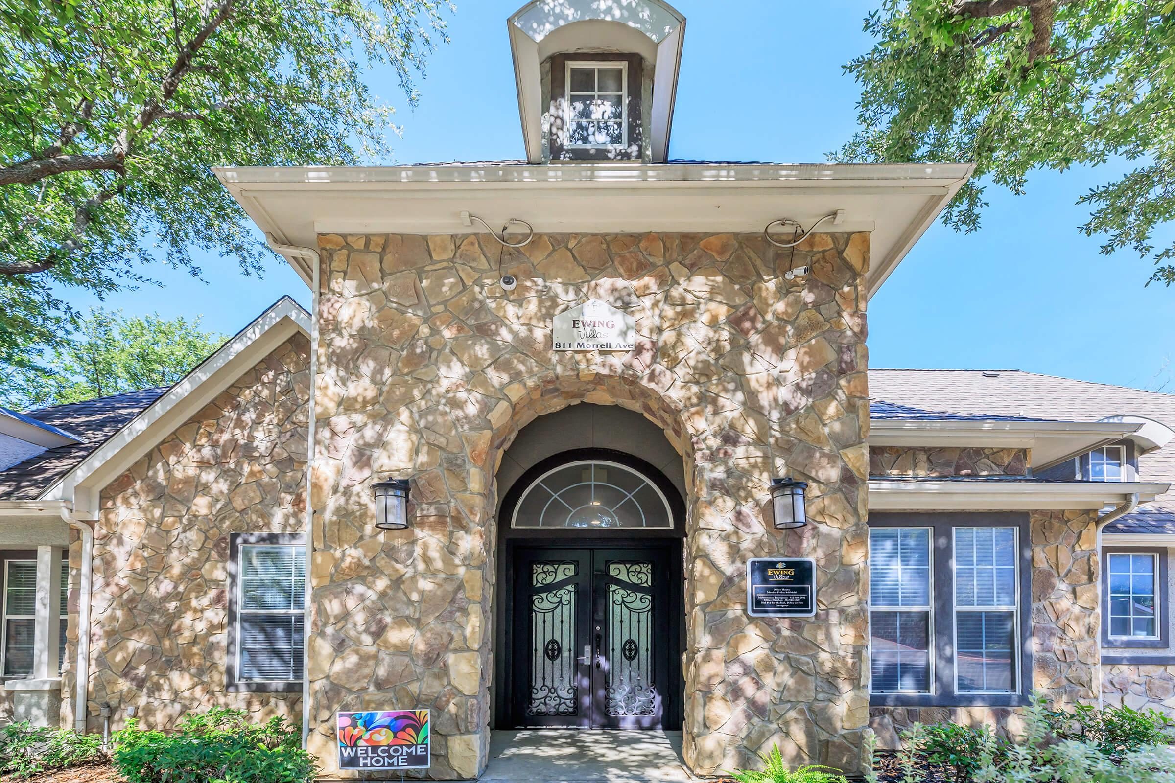 Front entrance of a stone-clad building featuring large arched doors, decorative lanterns, and a welcoming sign. Surrounded by trees and under a clear blue sky, the facade showcases a combination of traditional and modern architectural elements.