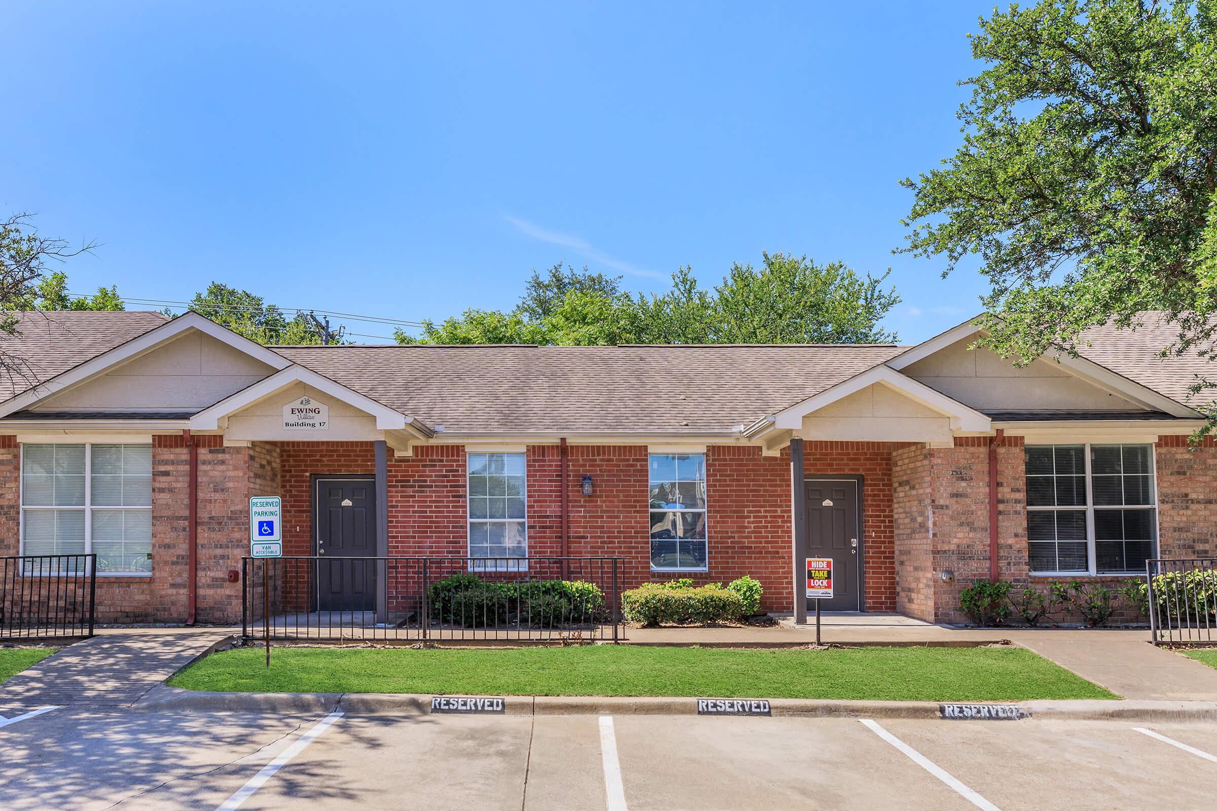 A row of brick apartments with a sidewalk and small lawn. Each unit has a front door and windows, with a sign by the entrance. The sky is clear and blue, and trees are visible in the background. The setting appears sunny and well-maintained, suitable for residential living.