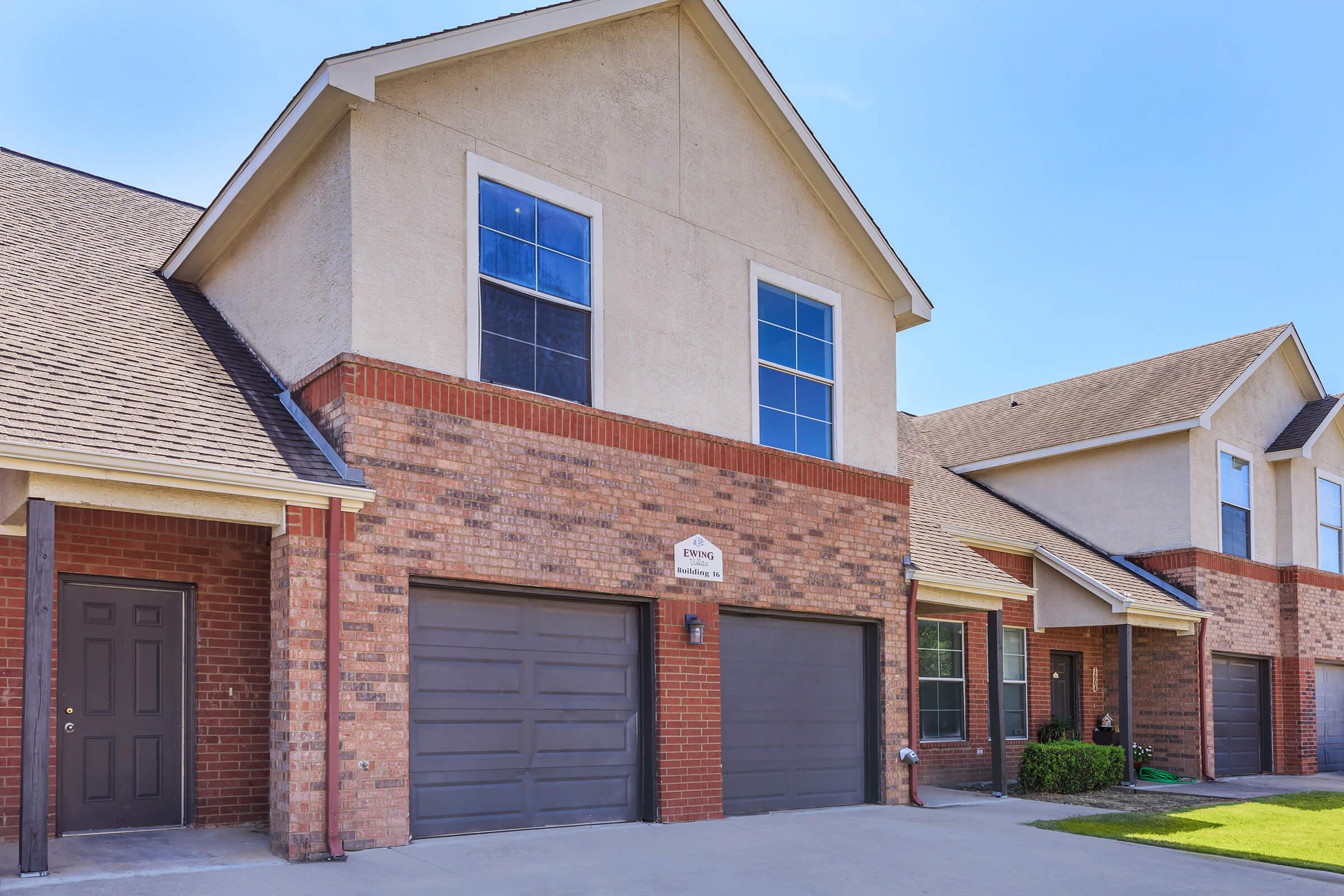 A row of two-story townhouses featuring brick exteriors and beige siding. Each unit has a garage with double doors, a front entrance, and large windows. The driveway is paved, and there is a well-maintained lawn in front of the buildings under a clear blue sky.