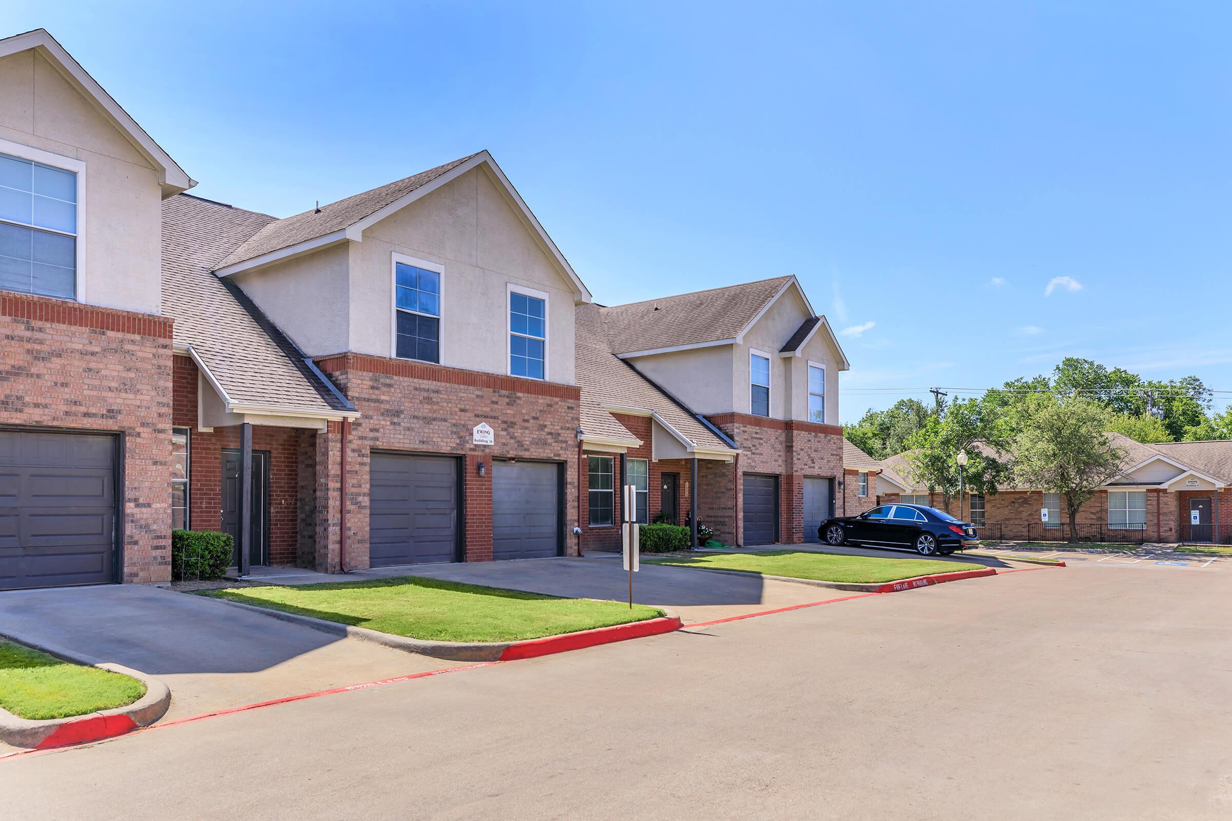A row of modern two-story townhouses with brick exteriors and manicured lawns. Each unit has a garage and a small porch. The scene features clear blue skies and well-maintained surroundings, suggesting a suburban residential area. A parked car is visible in the driveway.