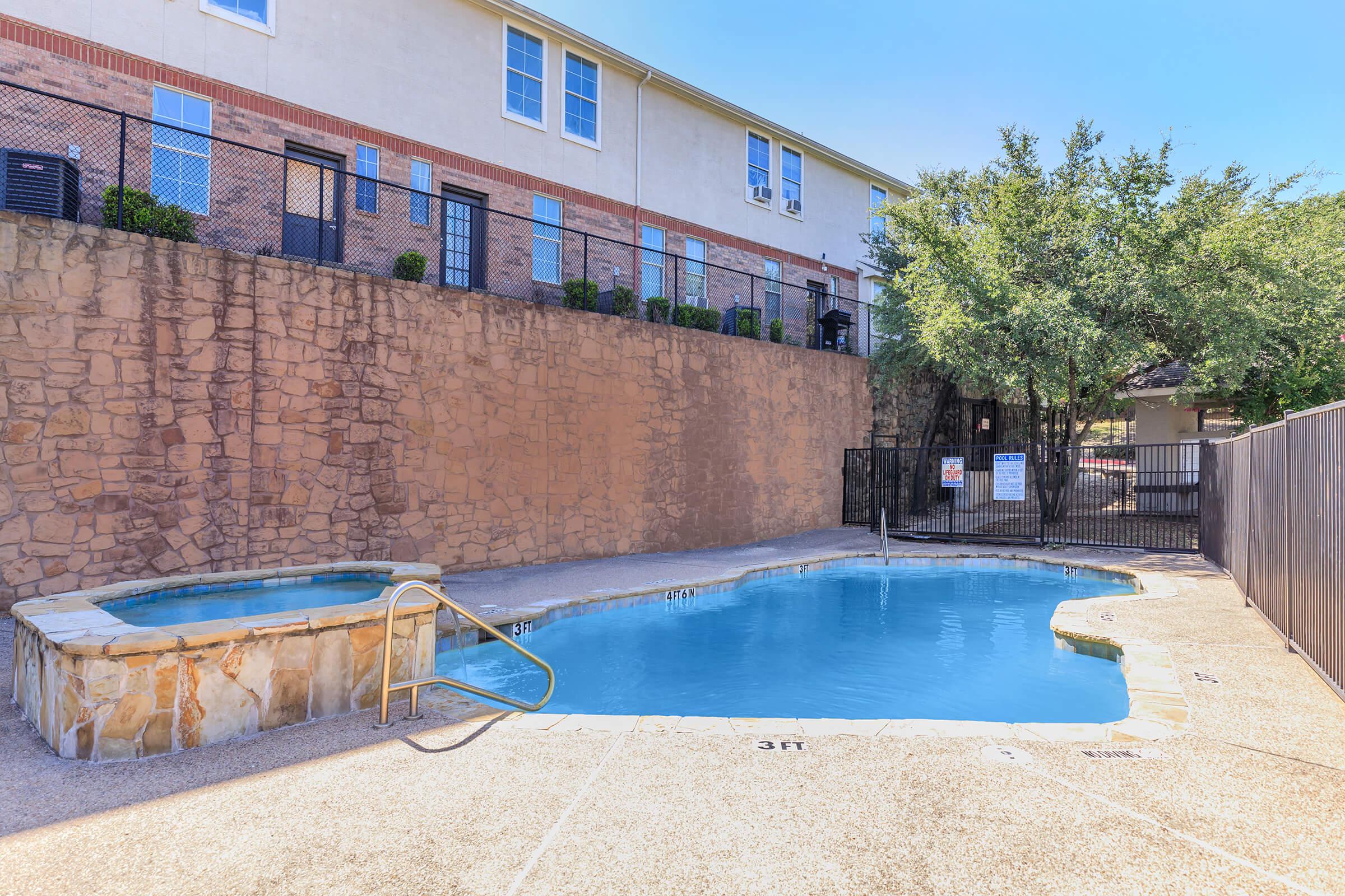 A clear blue swimming pool surrounded by a stone wall, with a smaller hot tub adjacent to it. The area is enclosed by a black fence, and there are trees nearby. In the background, a multi-story building can be seen. The pool has depth markers visible along the edge.