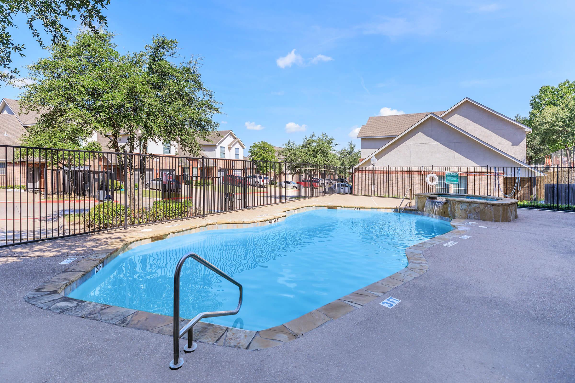 A clear swimming pool surrounded by a stone border, with a ladder at one end. Lush trees and residential buildings are visible in the background. The sky is bright blue with a few clouds, creating a warm and inviting atmosphere. A fenced area encloses the pool for safety.