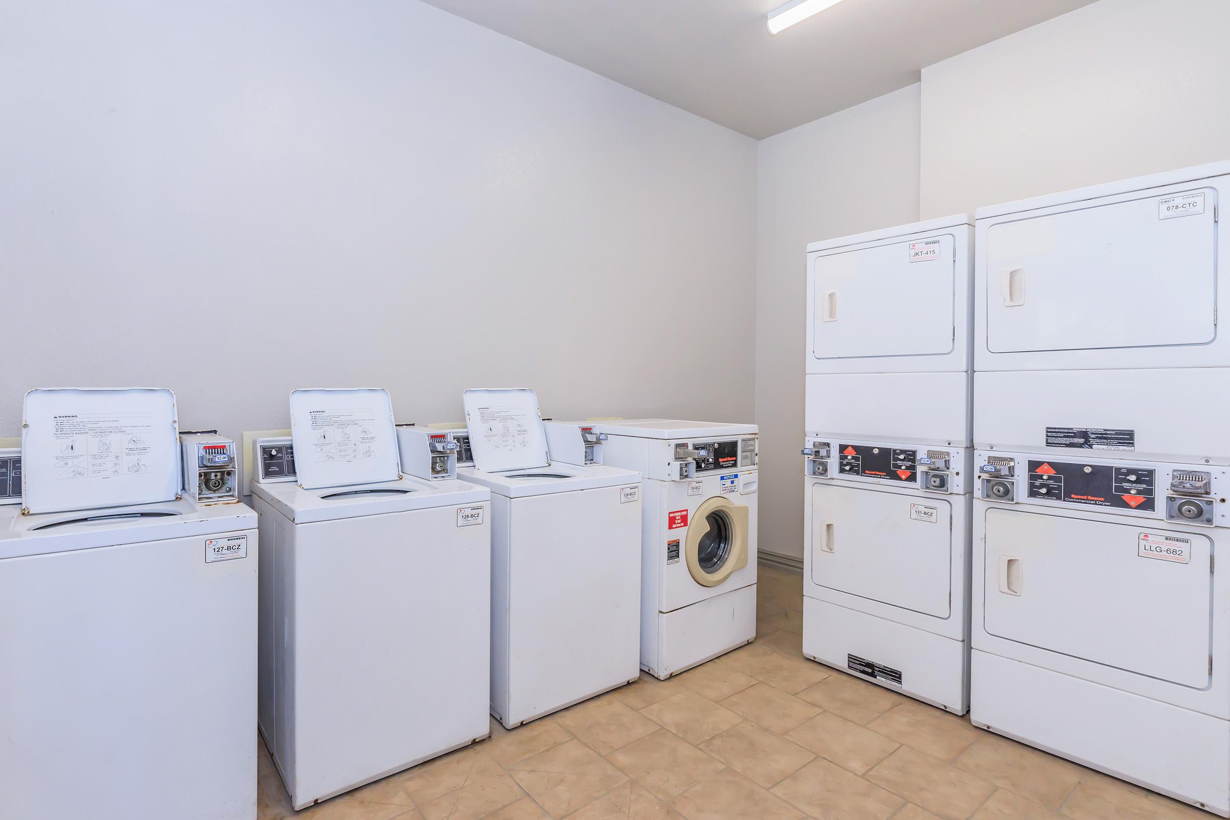 A laundry room featuring several white coin-operated washing machines and dryers. The machines are against a light-colored wall, with a tiled floor. The environment is clean and well-lit, offering a functional space for doing laundry.