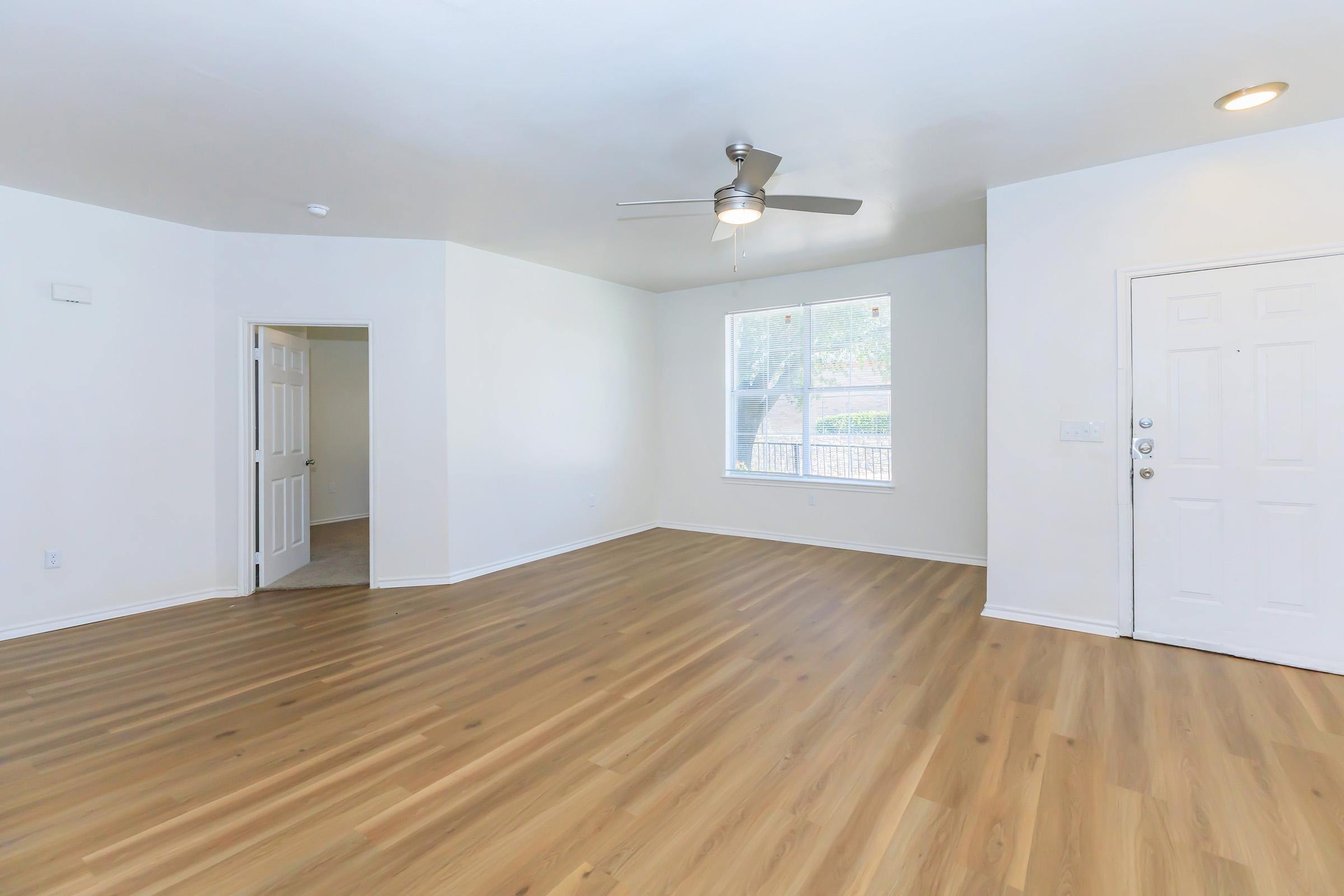 A spacious living room with light-colored walls and a ceiling fan. The floor is covered with light wood laminate. There is a large window allowing natural light and a view of the outside. An entrance door is visible on the right, and a door leading to another room is partially open in the background.