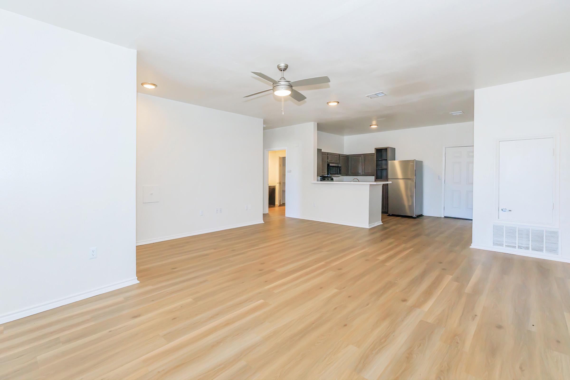 A spacious and bright living area with light-colored walls and wooden flooring. There is a ceiling fan installed, and the kitchen is partially visible in the background, featuring modern appliances and a bar counter. The overall atmosphere is clean and inviting, suitable for a contemporary home.