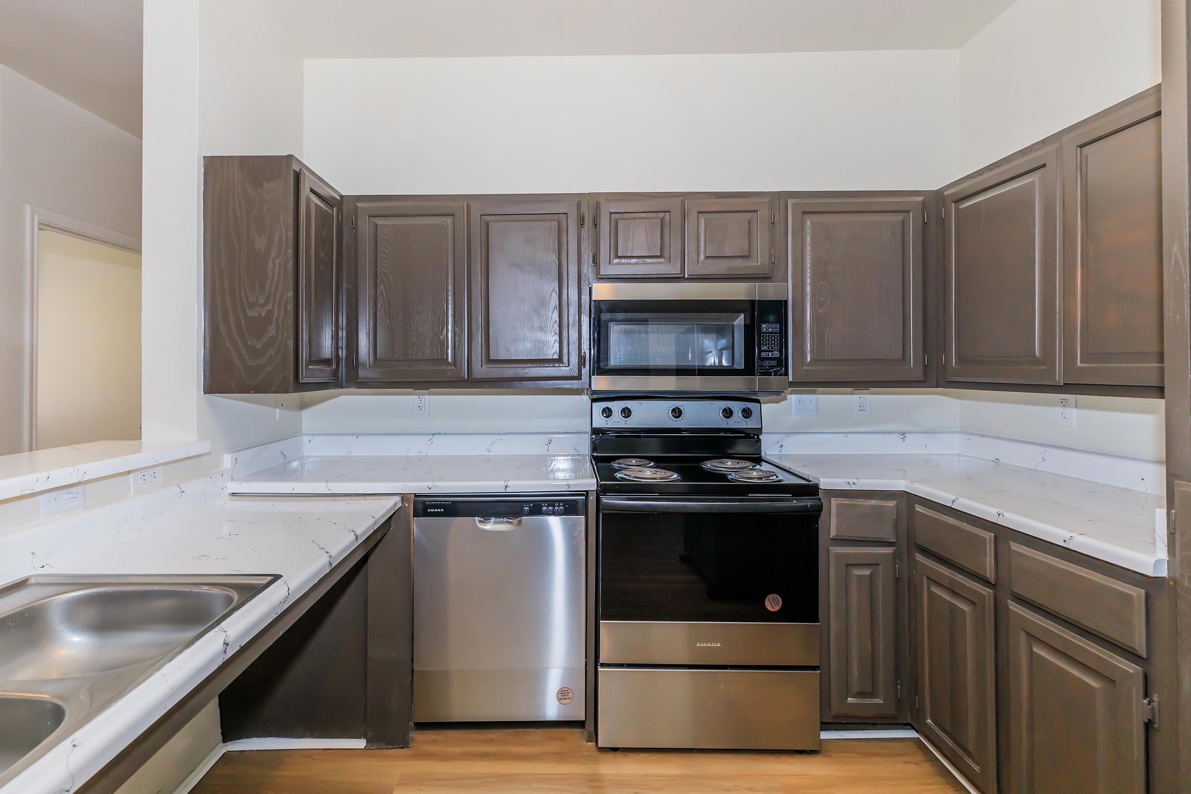Modern kitchen featuring dark wooden cabinetry, a black stove, and a stainless steel dishwasher. The countertops are light and the kitchen has a spacious layout with ample storage. The walls are painted white, creating a bright and clean atmosphere.