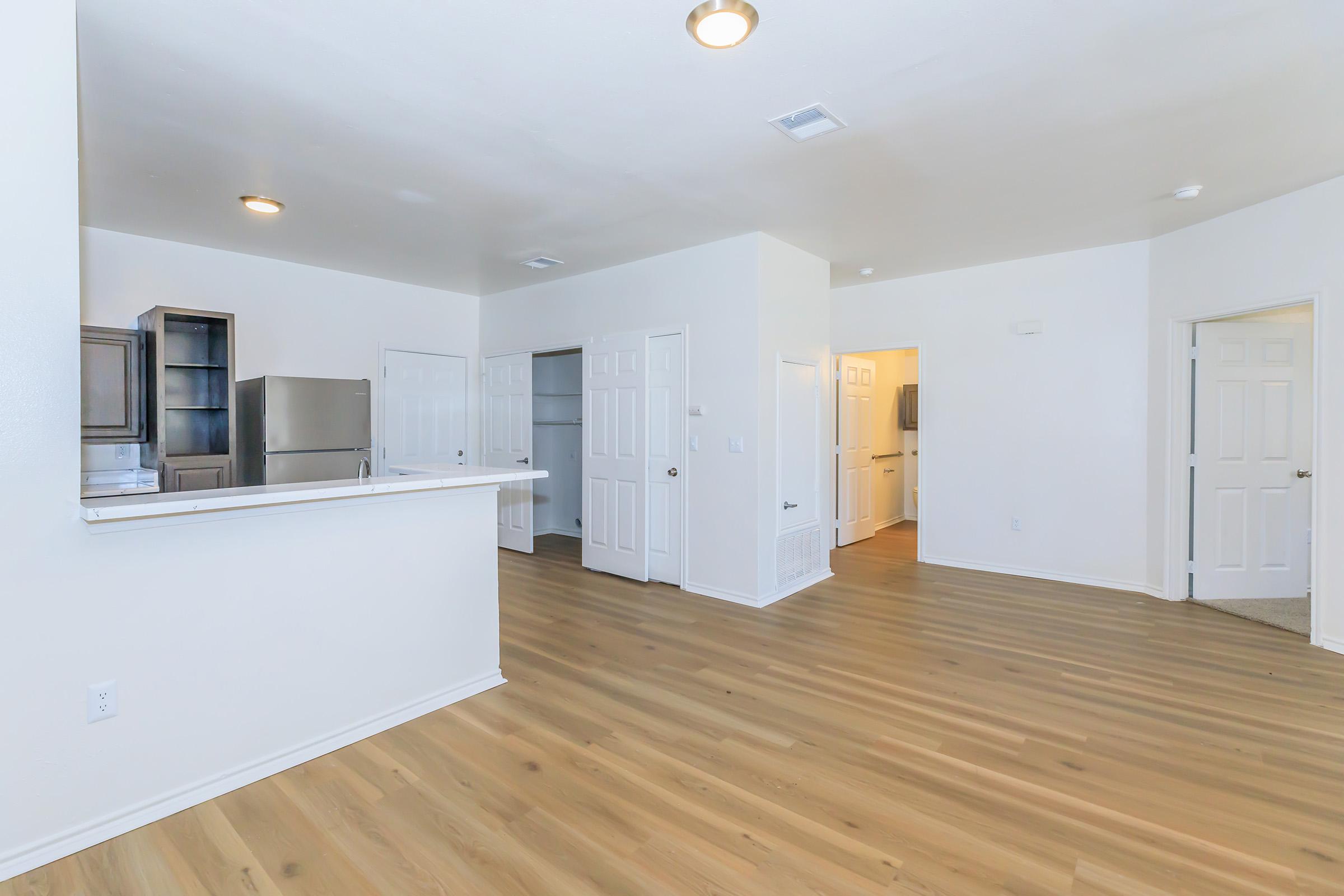 Interior view of a modern, spacious apartment with light-colored walls and wood-style flooring. The open layout features a kitchen bar area, a refrigerator visible in the background, and multiple closed doors leading to other rooms. Bright lighting from ceiling fixtures enhances the airy feel of the space.