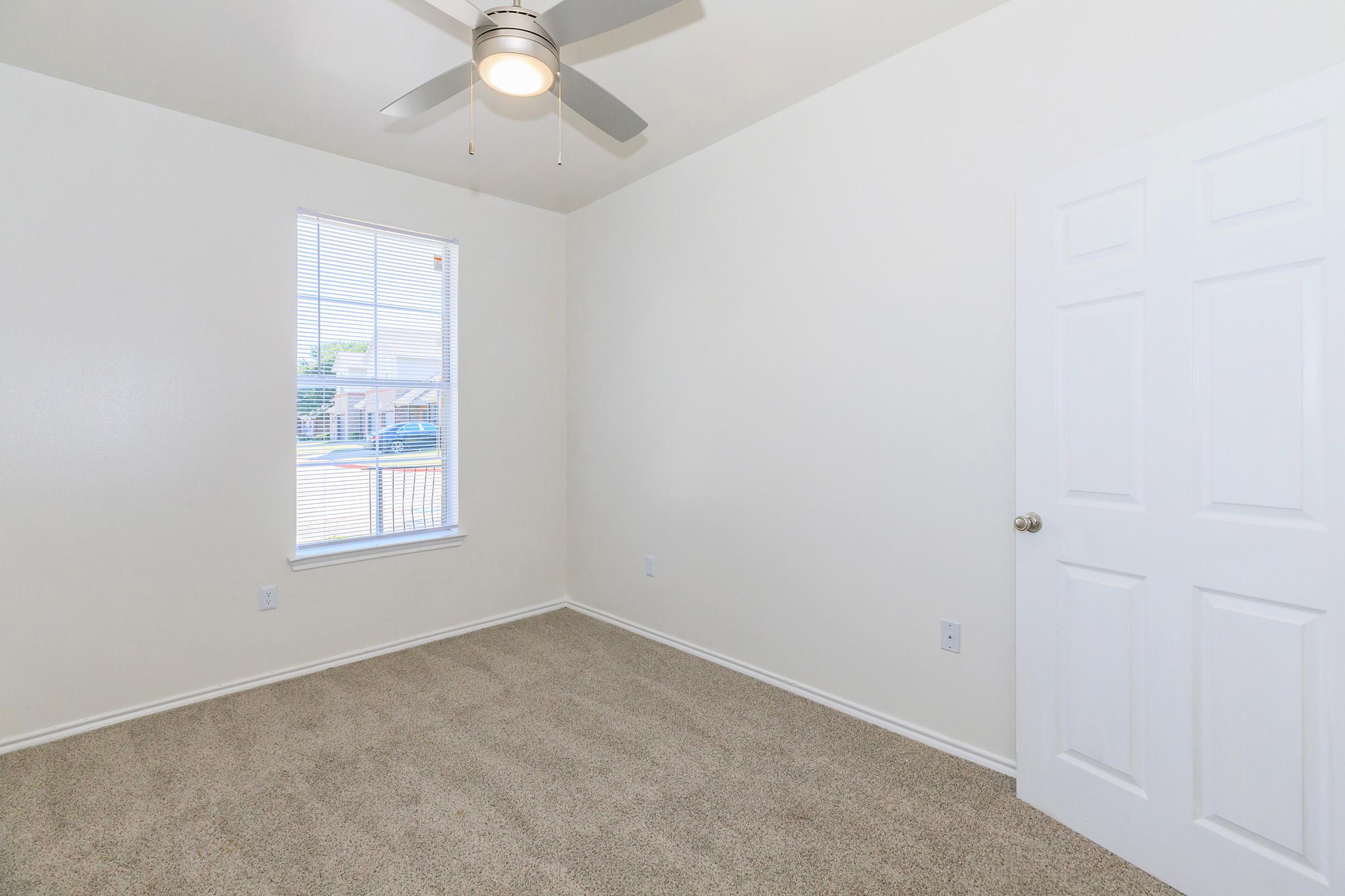 Empty room with light beige carpeting and white walls. A ceiling fan is mounted on the ceiling, and there is a window with blinds allowing natural light in. A closed white door is visible on the right side of the image. The overall atmosphere is clean and spacious, suitable for various purposes.