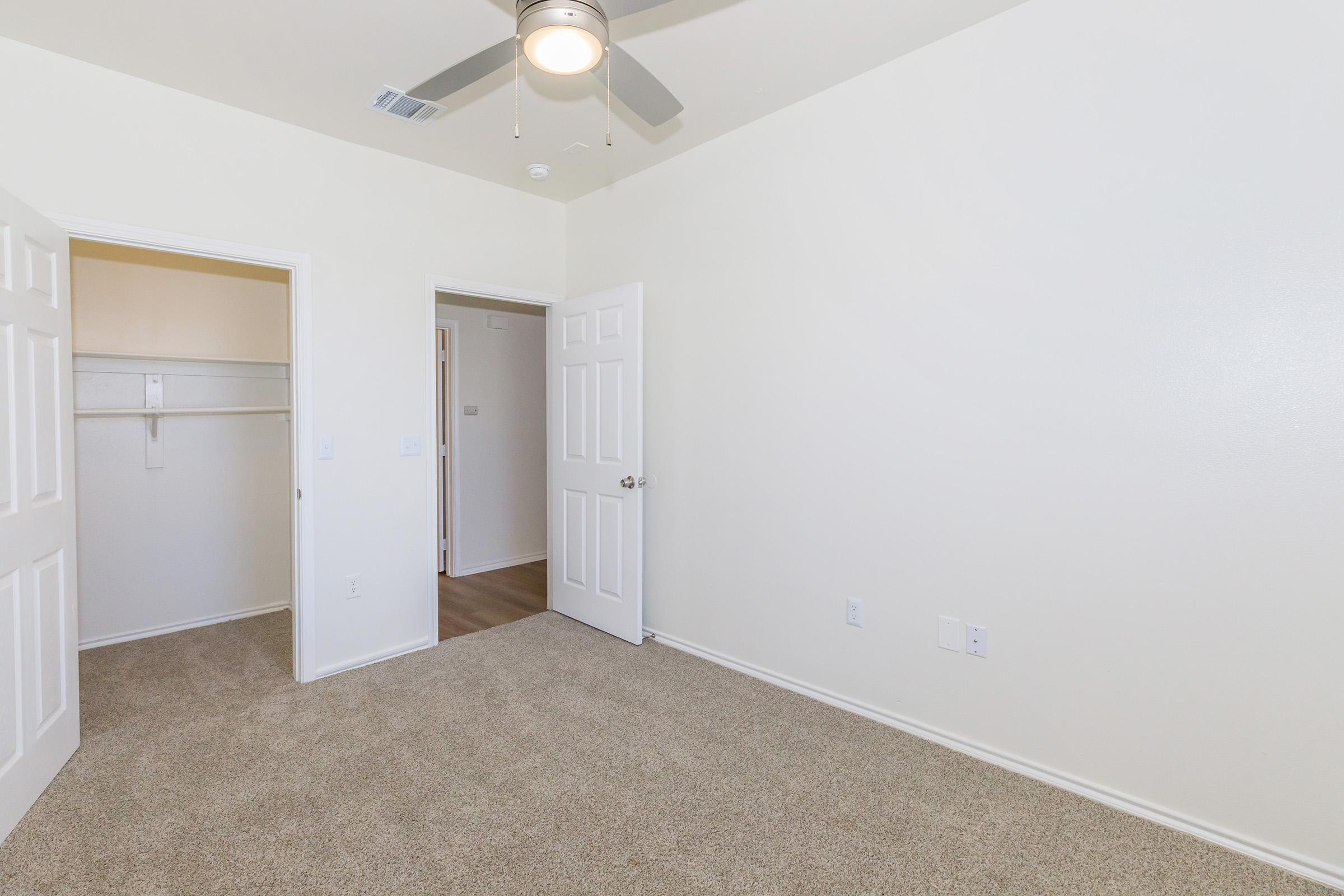 A plain, empty room with light beige carpet and white walls. Two closed doors are visible, one leading to a closet with shelves. A ceiling fan is mounted above, and the room has a bright and airy feel with natural light.
