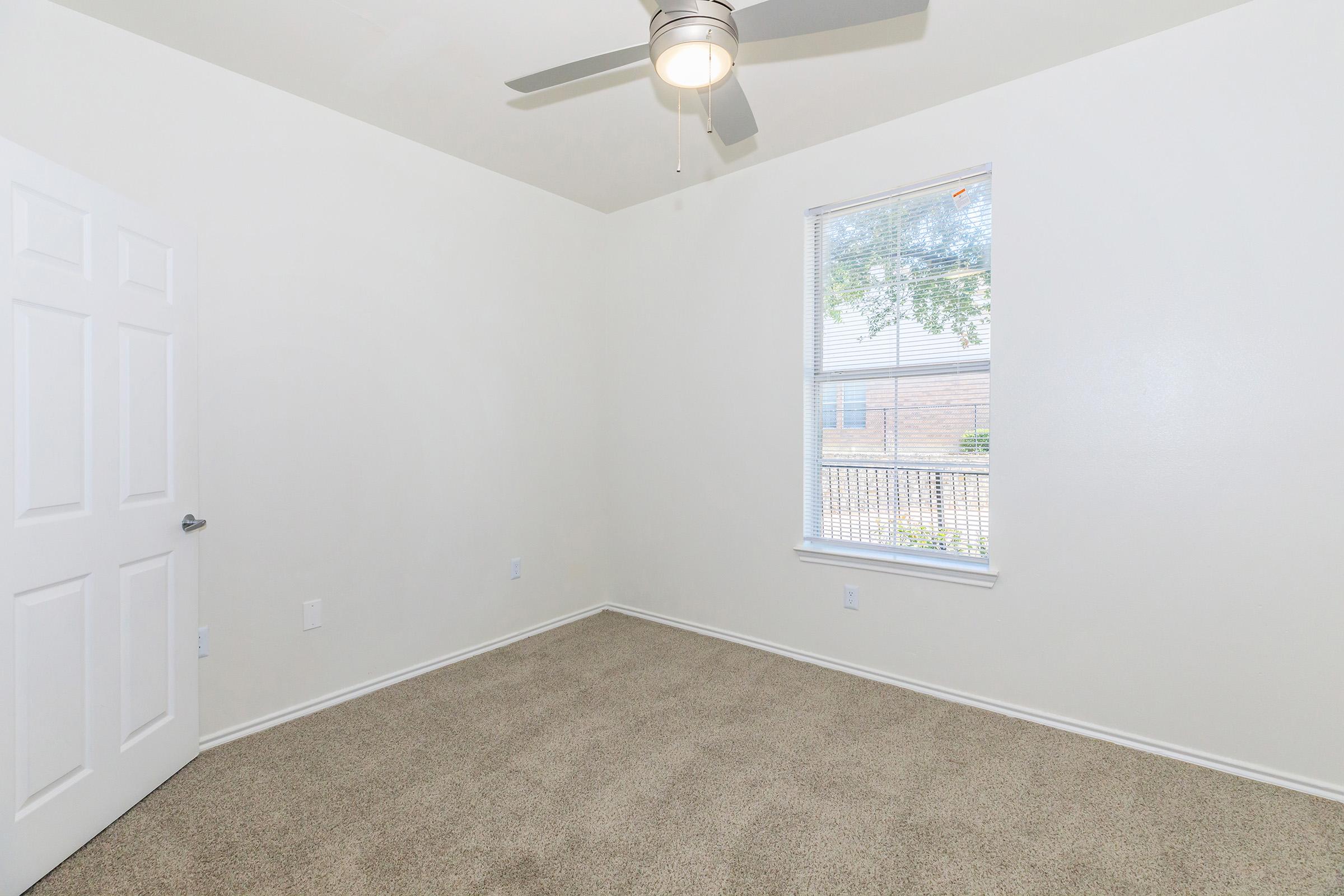 A vacant room with light-colored walls and a ceiling fan. There is a window with a view of the outdoors, letting in natural light, and a closed white door on one side. The floor is covered in tan carpet, creating a cozy atmosphere.