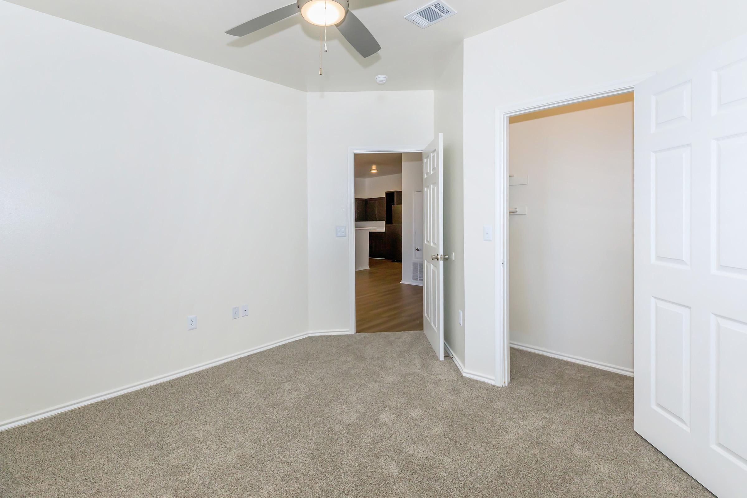Empty room with beige walls and carpeted floor. A ceiling fan hangs from the ceiling. There are open double doors leading to a closet on one side and an entrance to another room on the opposite side. Natural light comes from the doorway, illuminating the space.
