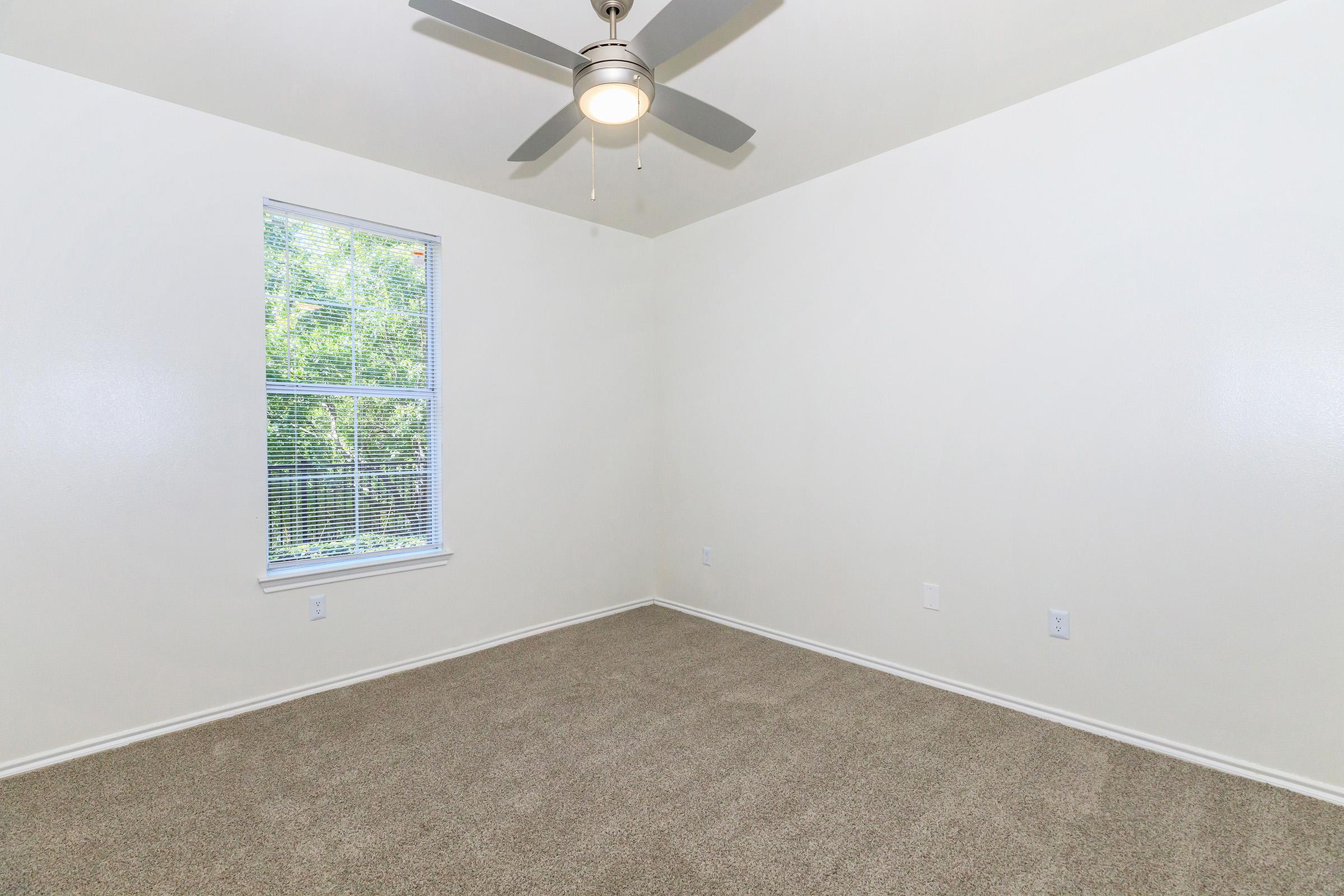 Empty room with light beige walls and a ceiling fan. A window with blinds allows natural light to enter, offering a view of greenery outside. The floor is covered with light brown carpet, creating a warm and inviting atmosphere. The room appears spacious with minimal furnishings.