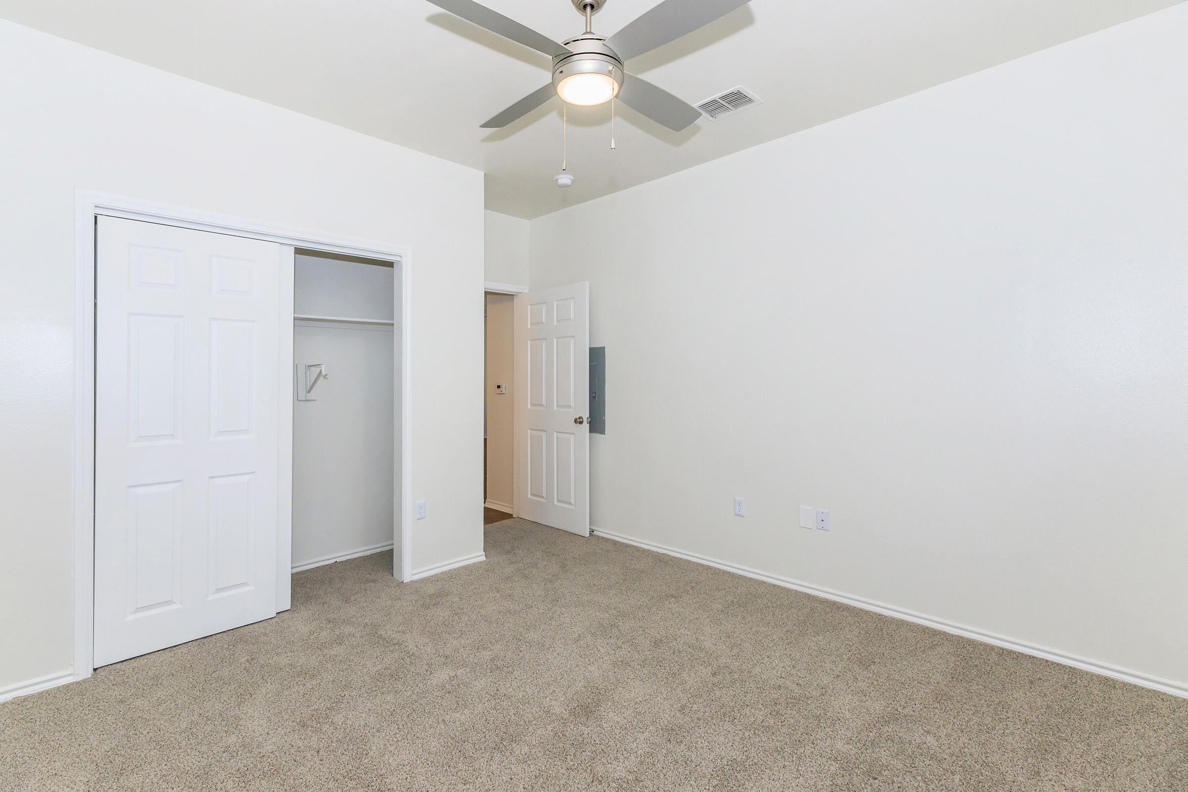 A simple, well-lit bedroom featuring neutral-colored walls and carpeting. It includes a ceiling fan, a closet with double doors on one side, and an open door leading to another room. The space is empty, providing a blank canvas for potential decoration.