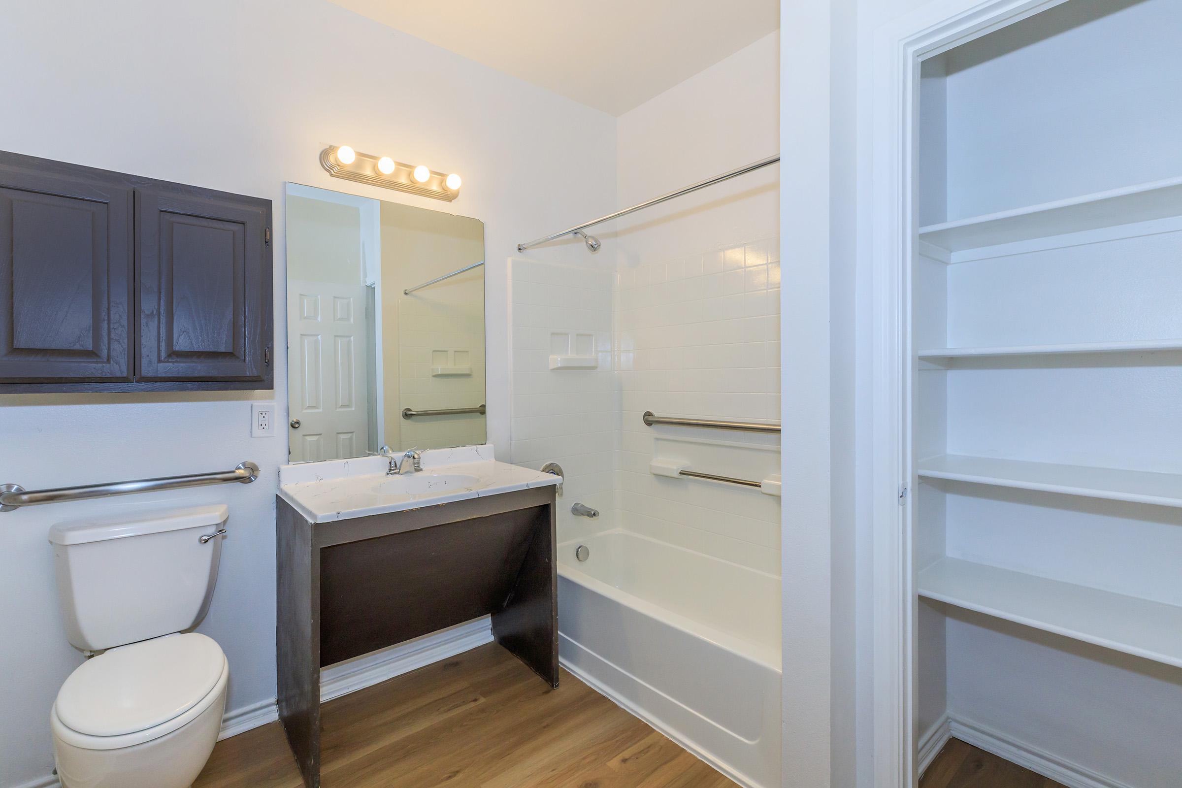 A modern bathroom featuring a white bathtub with a shower, a rectangular sink with cabinetry, a wall-mounted mirror with lighting, a white toilet, and a shelving unit. The walls are painted light colors, and the flooring is wooden.