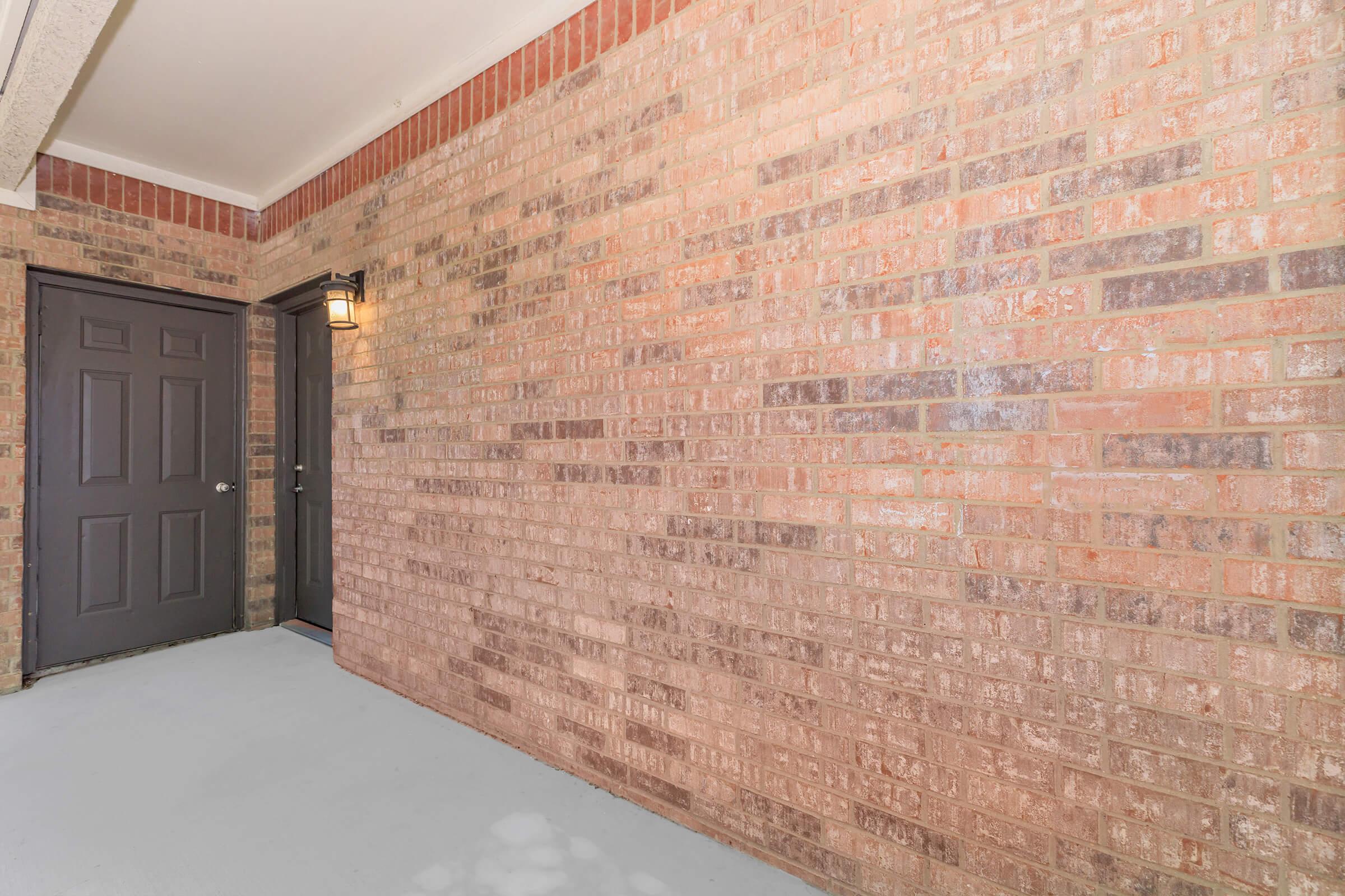 Exterior view of a brick wall with a gray door on the left side. A light fixture is mounted next to the door, illuminating the entrance area. The floor is a light-colored surface, and the overall space appears clean and well-maintained, suitable for residential entry.