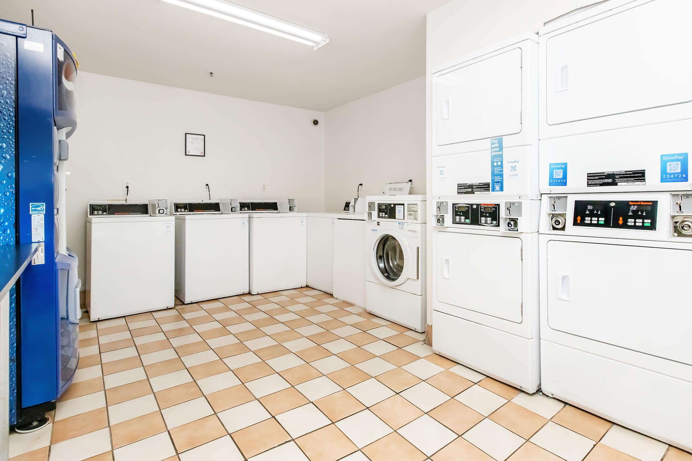 A clean and bright laundry room featuring several white washing machines and dryers arranged against the wall. The floor is tiled with beige and white squares. A blue vending machine is visible in the corner, and there's a wall with a framed notice or schedule. Good lighting enhances the spacious atmosphere.