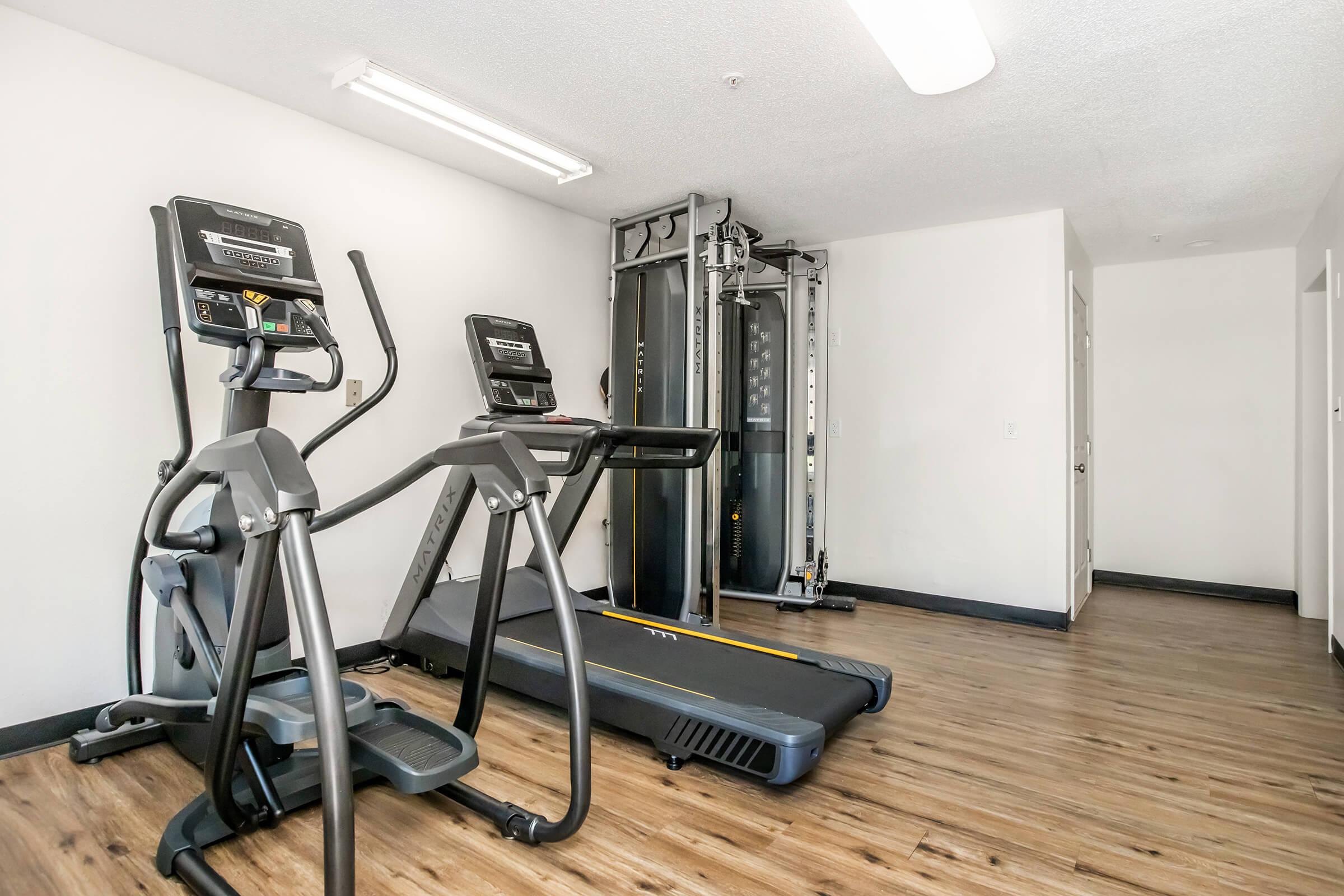 A bright, modern gym space featuring two cardio machines (an elliptical and a treadmill) and a multi-station weight machine. The flooring is a light wood, and the walls are painted white, creating an open and inviting atmosphere. There's ample space for workouts with good lighting.