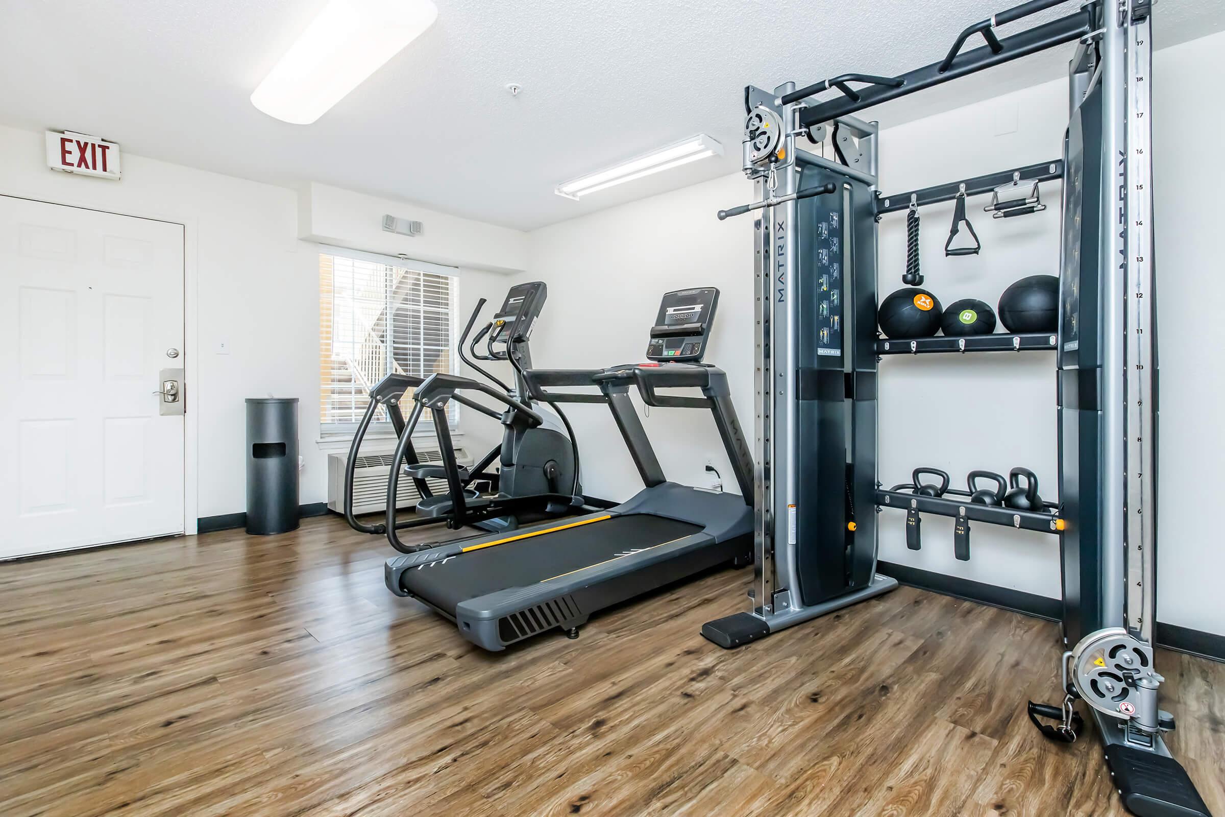 A modern gym interior featuring a treadmill, an elliptical machine, and a multi-gym weight machine. The space includes fitness equipment such as exercise balls and weights, with wood-like flooring and bright lighting. Large windows provide natural light, and there is an exit door visible.
