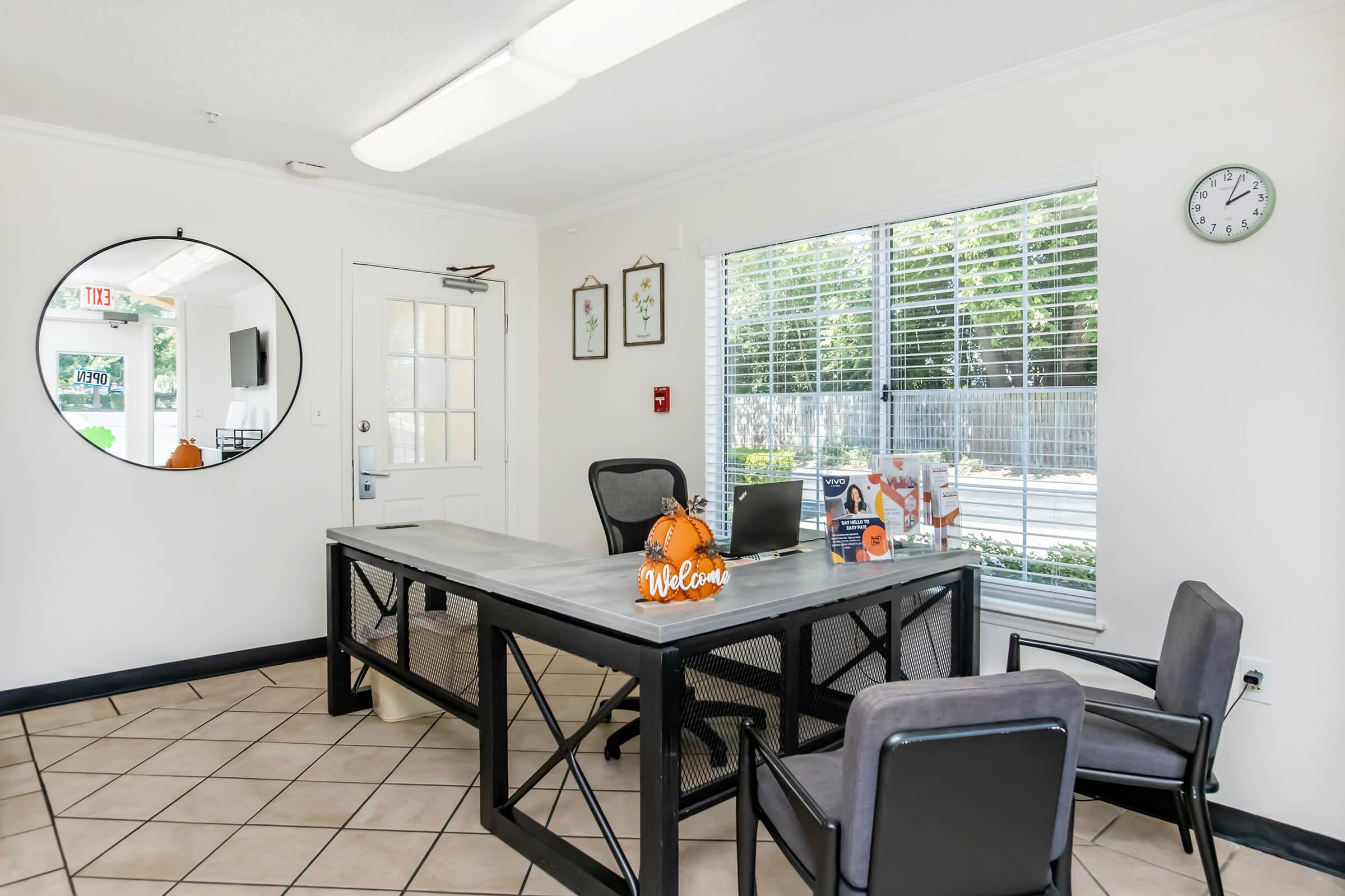 A welcoming office space featuring a modern desk with a "Welcome" sign, two black chairs, a large circular mirror on the wall, and a window with vertical blinds. A small clock is visible, and decor includes framed pictures and a seasonal pumpkin display. The overall setting is bright and organized.