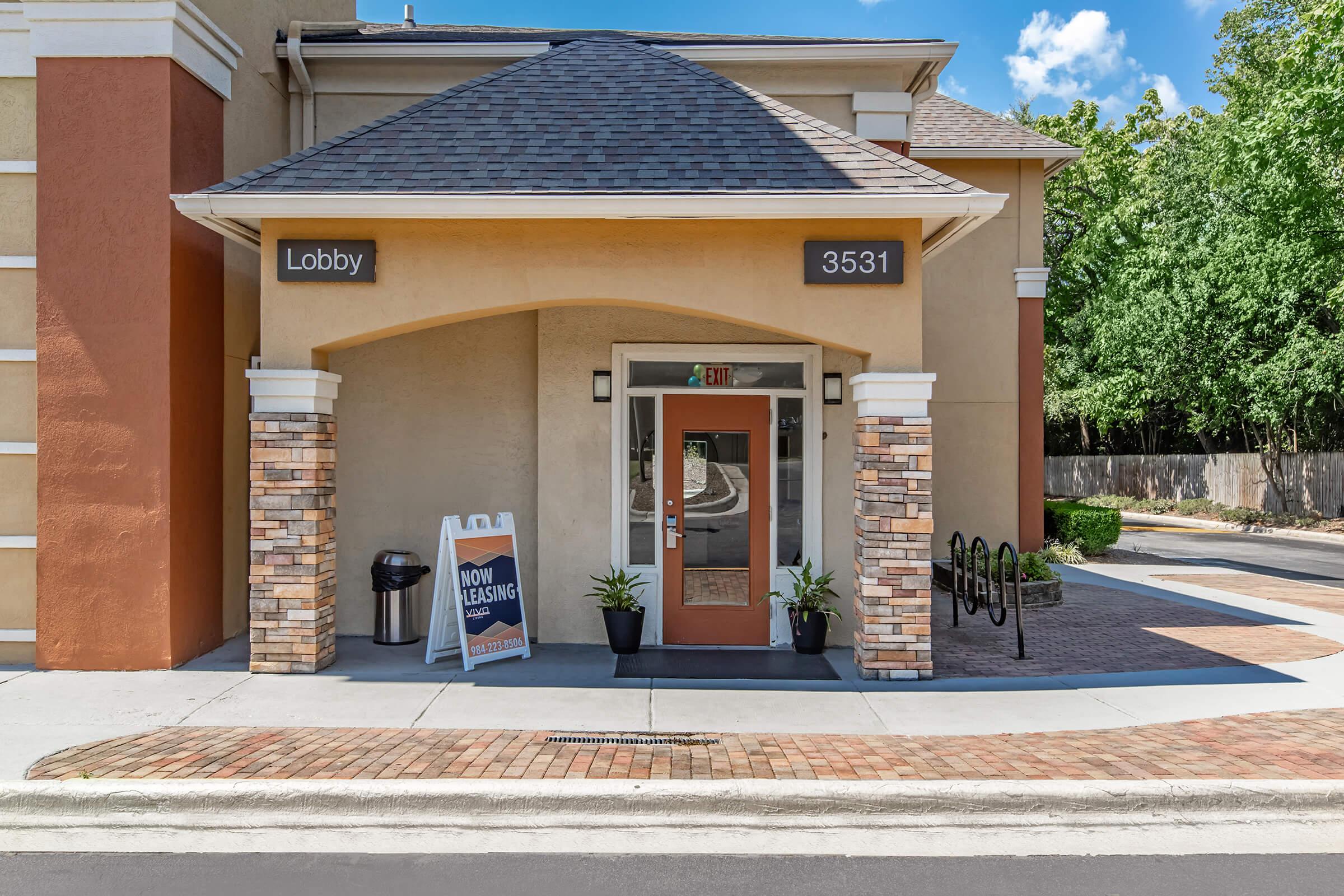 Entrance to a lobby with a modern design, featuring a wooden door, brick accents, and signs indicating "Lobby" and "Now Leasing." Two potted plants are flanking the door, and a bike rack is visible nearby. Clear blue skies and greenery are seen in the background.