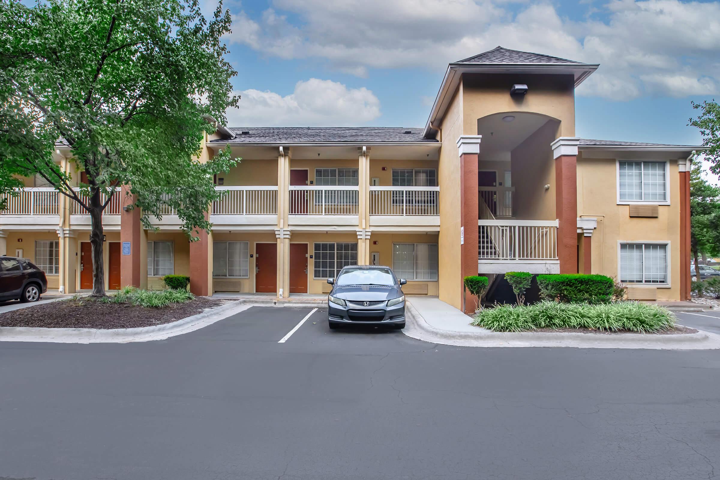 A two-story motel building with a cream-colored façade and orange accents, featuring multiple balconies. In the foreground, a parked black car is visible on a paved driveway, bordered by green shrubs and trees. The sky is partly cloudy.