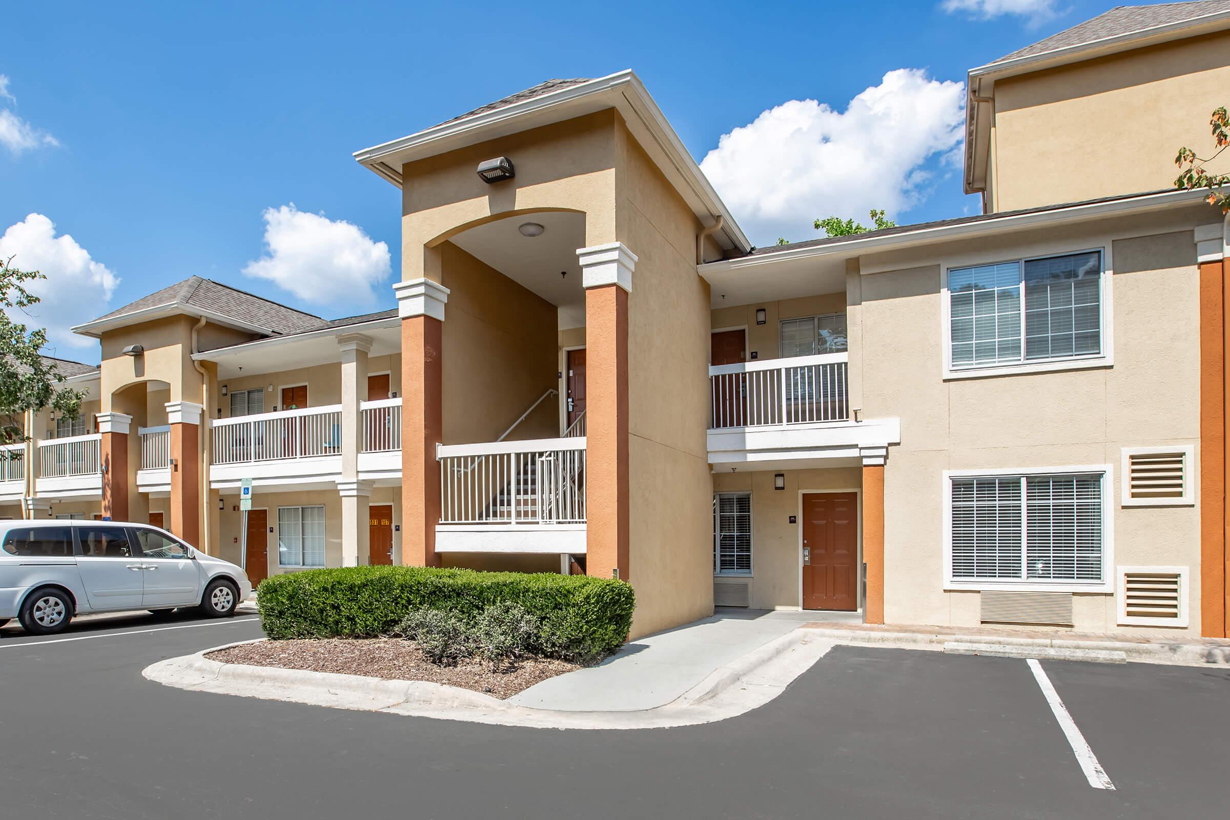A well-maintained, two-story apartment building with beige and orange accents. The building features balconies and several windows. In the foreground, a paved parking lot with a white car and neatly trimmed bushes adds to the tidy appearance. The sky is bright with scattered clouds.