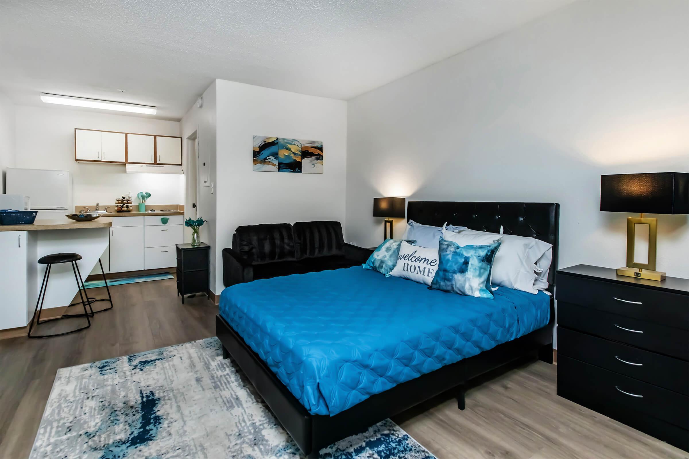 Cozy studio apartment featuring a queen-sized bed with blue bedding, a black sofa, and a stylish area rug. The kitchen area is visible in the background, equipped with appliances and cabinets. Soft lighting from bedside lamps creates a welcoming atmosphere.