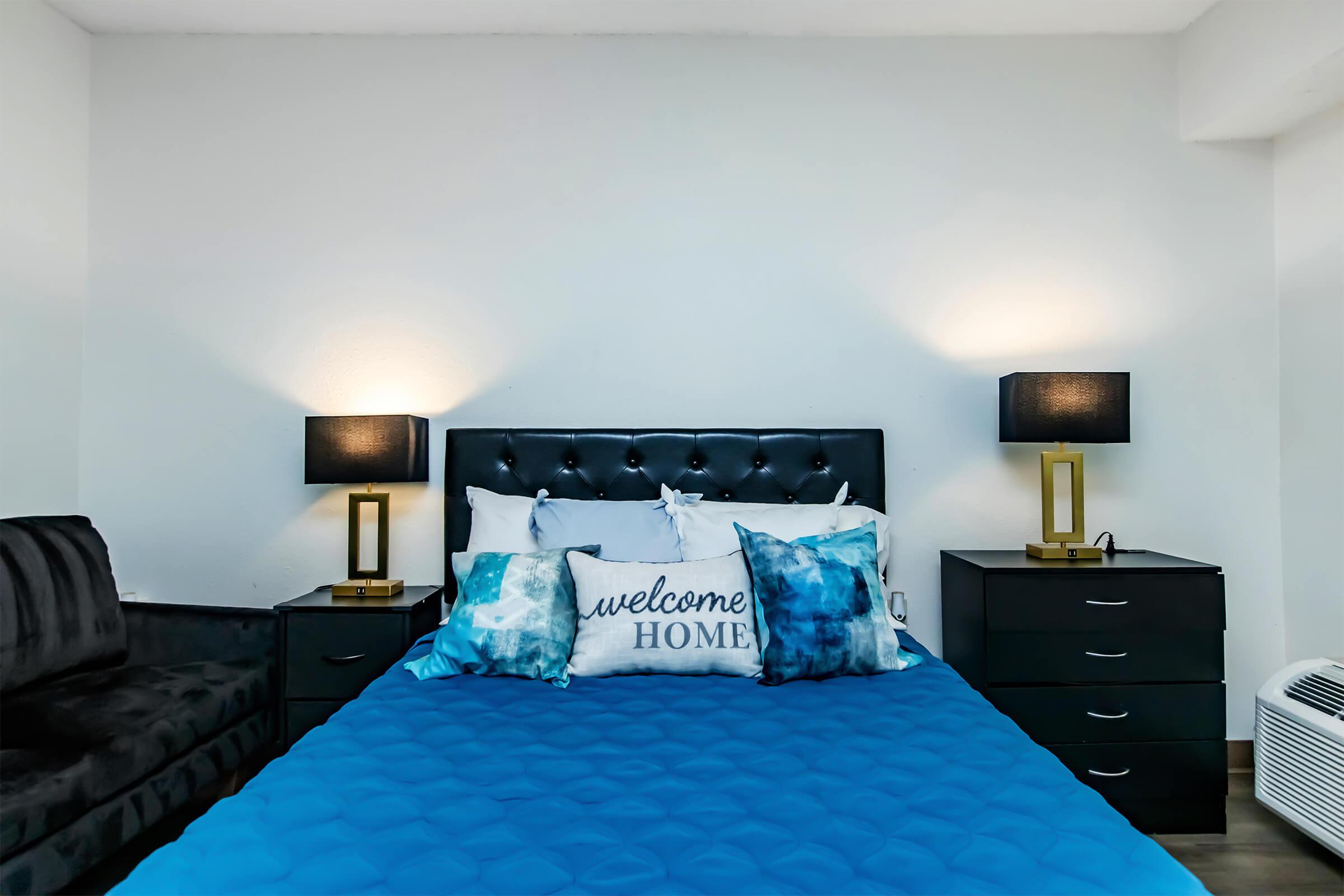 A cozy bedroom featuring a blue quilted bedspread and decorative pillows, including one that says "welcome HOME." Two stylish bedside lamps with black shades flank a dark dresser. A plush black sofa is visible in the corner, and the walls are painted white to create a bright, welcoming atmosphere.
