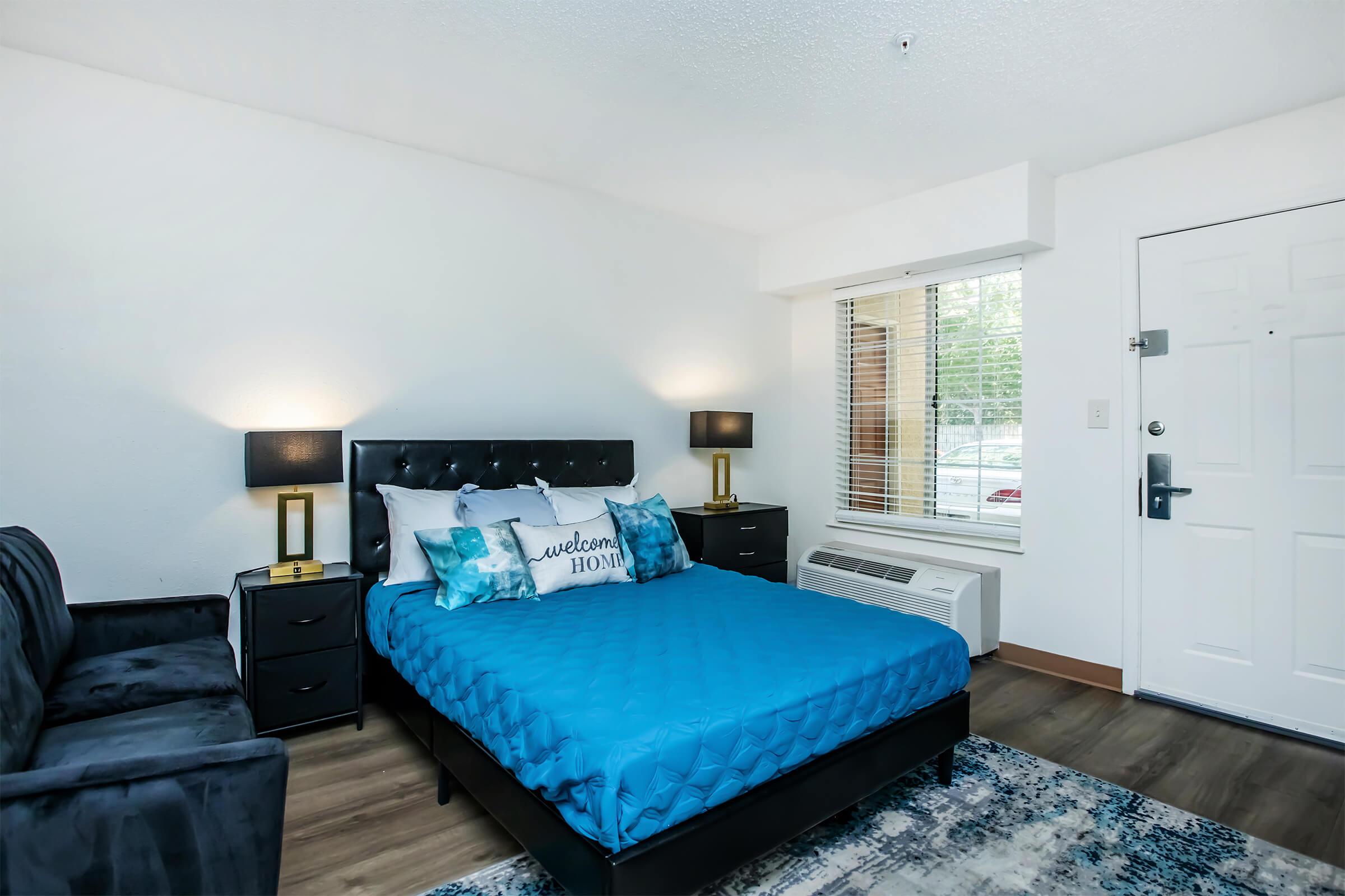 A cozy bedroom featuring a queen-sized bed with a blue quilt and decorative pillows, a black upholstered headboard, two bedside lamps, a black armchair, and a small window letting in natural light. The room has wooden flooring and a white door leading outside.
