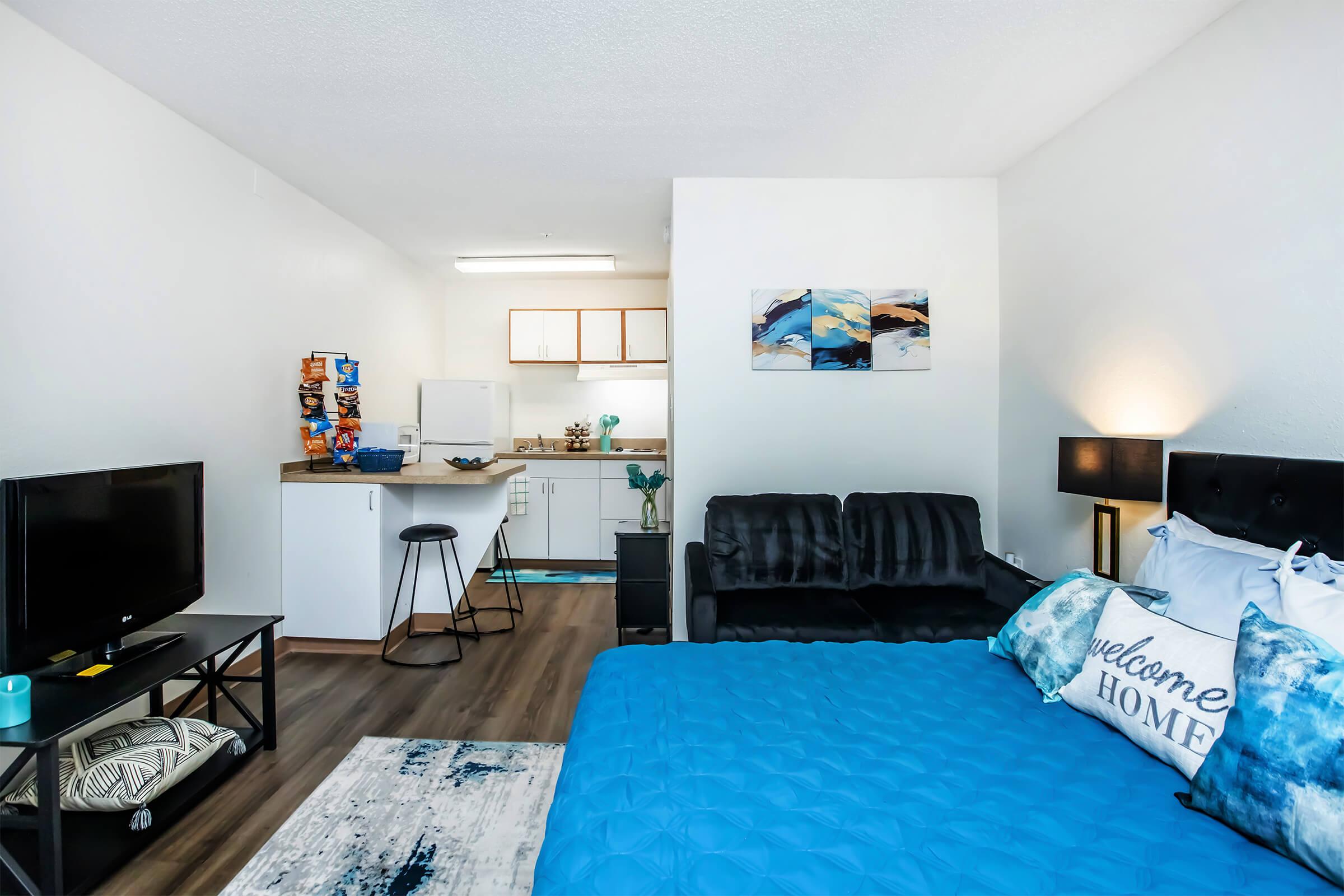A cozy studio apartment featuring a bed with blue bedding, a black sofa, and a television. The kitchen area includes a refrigerator and cabinets, while a painting hangs on the wall. There’s a welcome home pillow and a small dining area with a bar stool, creating a warm and inviting ambiance.